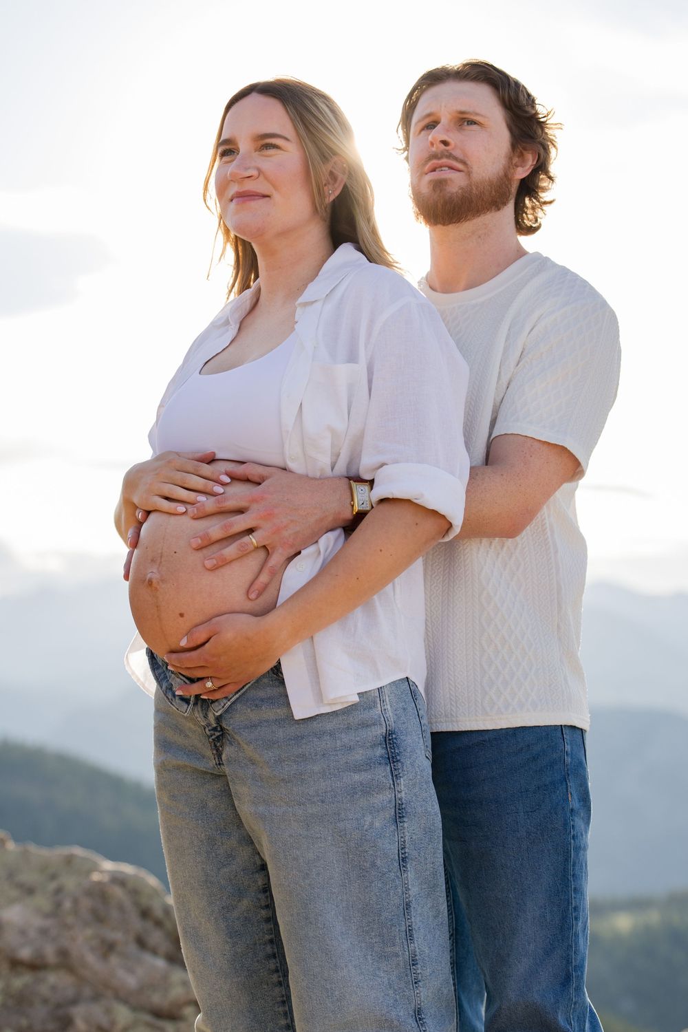 Pregnant couple standing outdoors with mountain views, man embracing woman's baby bump, both wearing casual white tops and jeans.