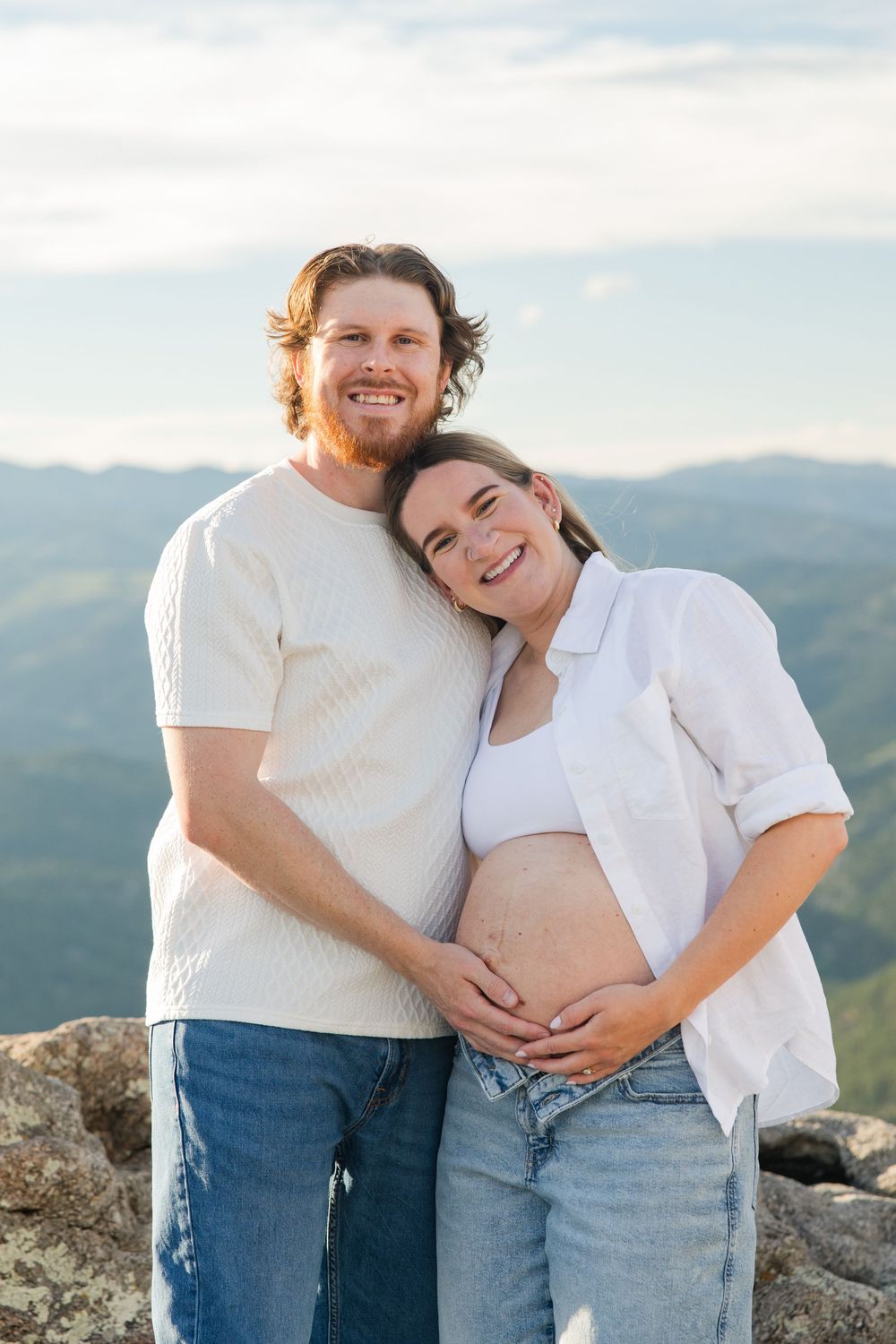 Smiling couple posing on mountain summit, pregnant woman in white shirt cradling baby bump with partner.