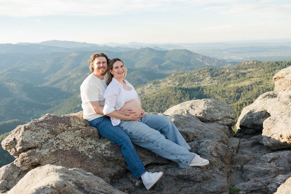 Couple sitting on rocky mountain summit, man embracing pregnant woman, scenic mountain valley backdrop, maternity photo session.