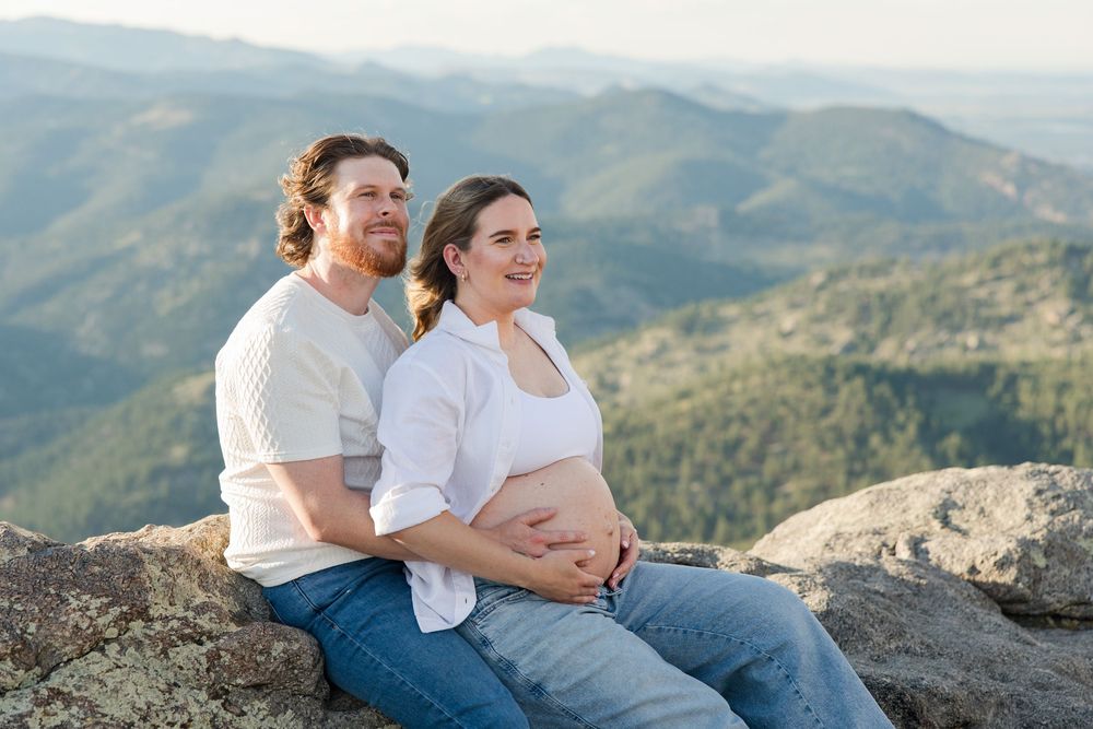 Expectant couple sitting on rocky mountain overlook, embracing and gazing at scenic valley views.