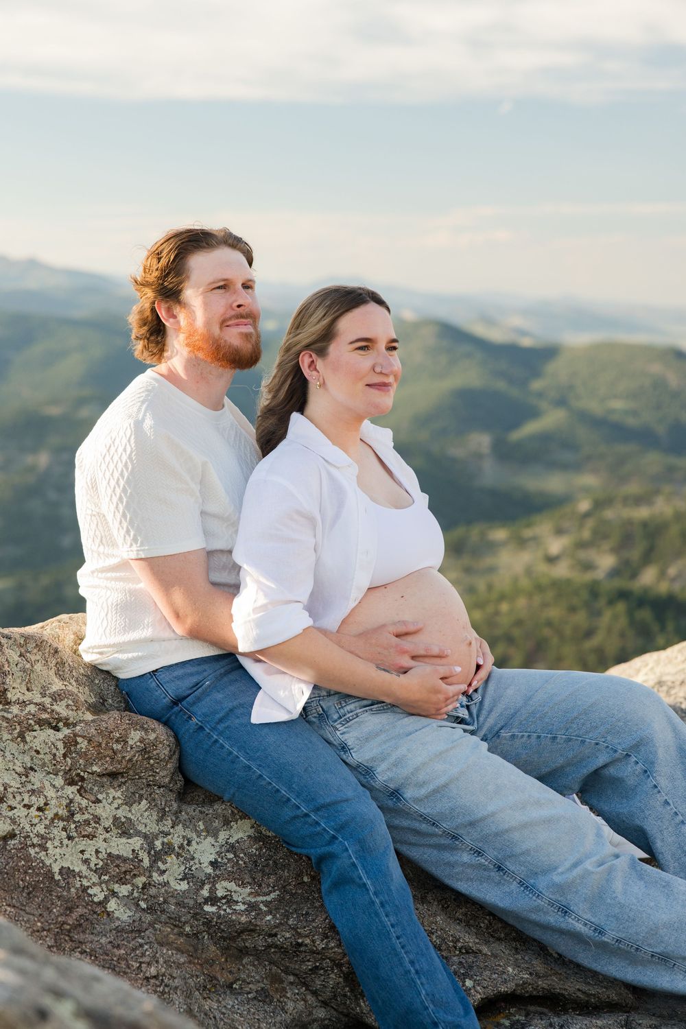 Pregnant couple in white shirts and jeans sitting on a rocky overlook with mountain views, sharing a tender moment.