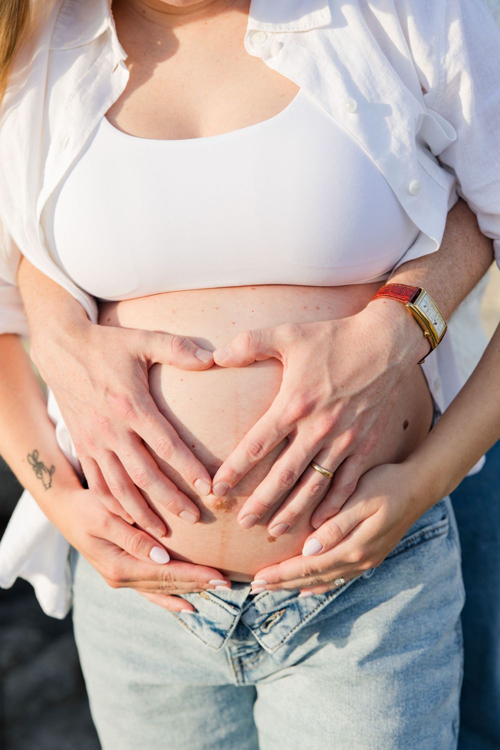 Pregnant woman in white crop top and jeans with two pairs of hands forming a heart shape on her baby bump.