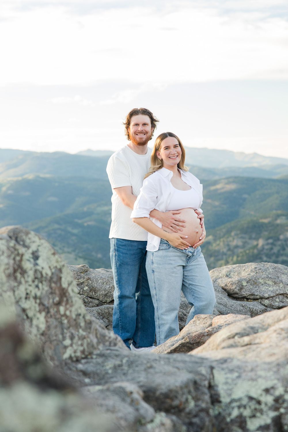 Smiling couple poses on mountain rocks, woman showing pregnant belly, scenic Colorado mountain range backdrop.
