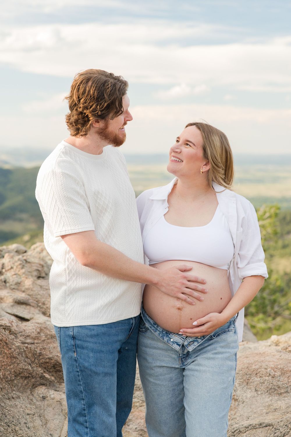 Smiling pregnant couple in white tops and jeans embrace on rocky overlook with scenic valley views.