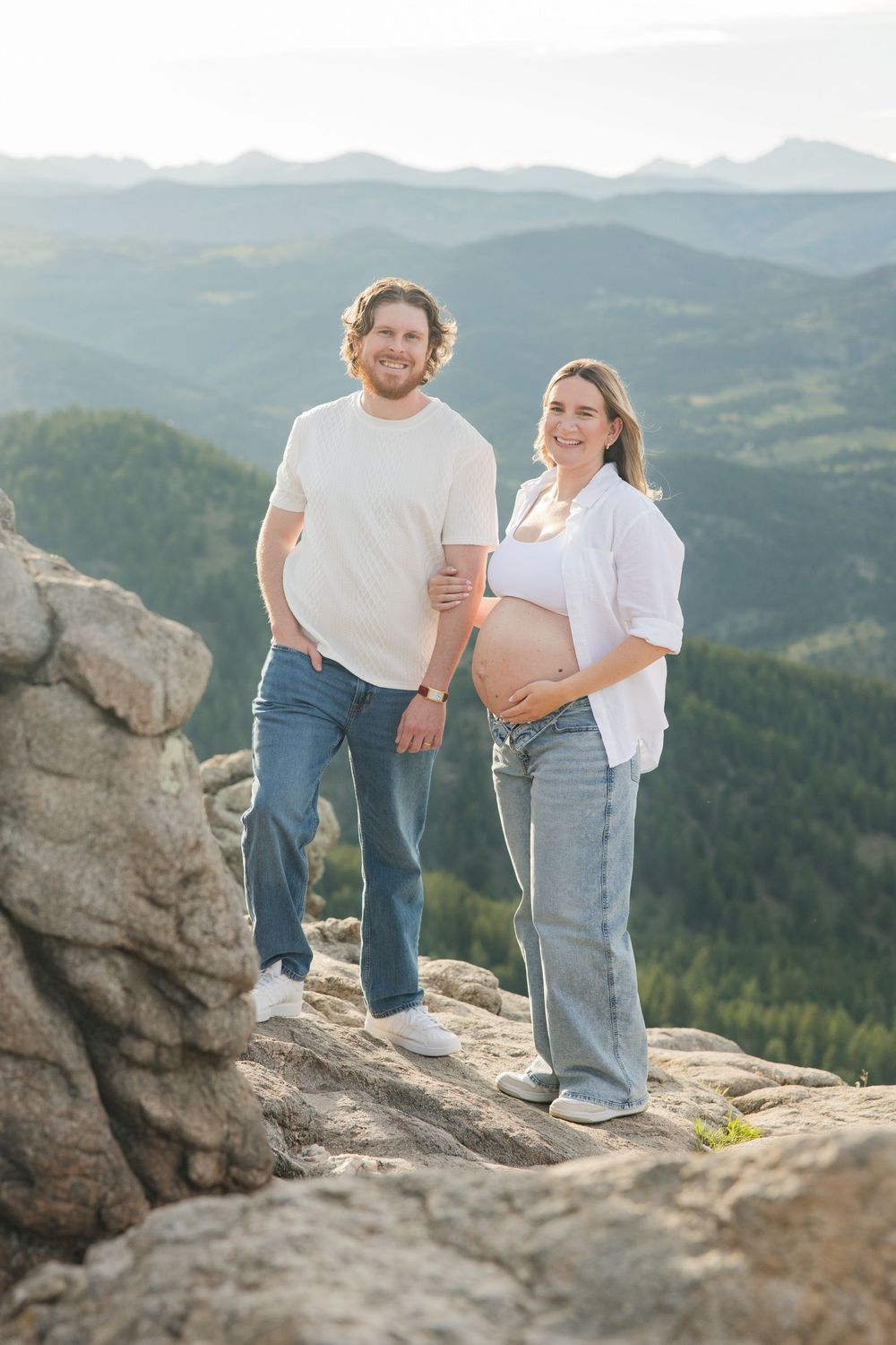 Happy couple posing on mountain rocks, pregnant woman showing baby bump, scenic green valley and mountains in background.