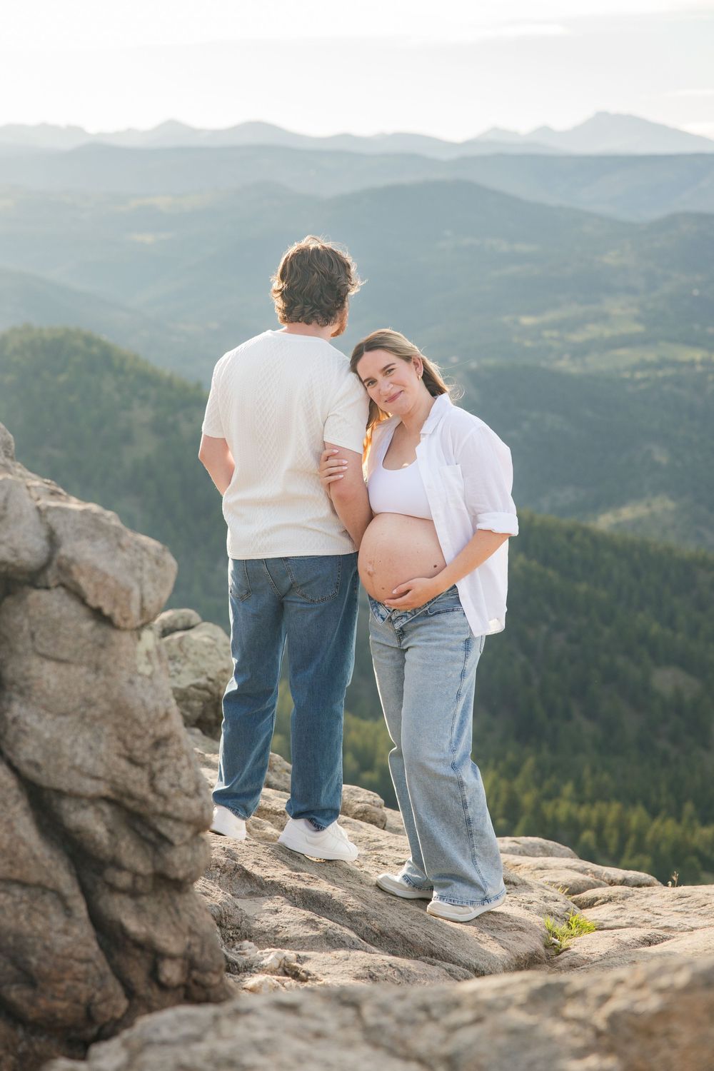 Pregnant woman in white shirt and jeans leans on partner at mountain overlook with scenic valley views.