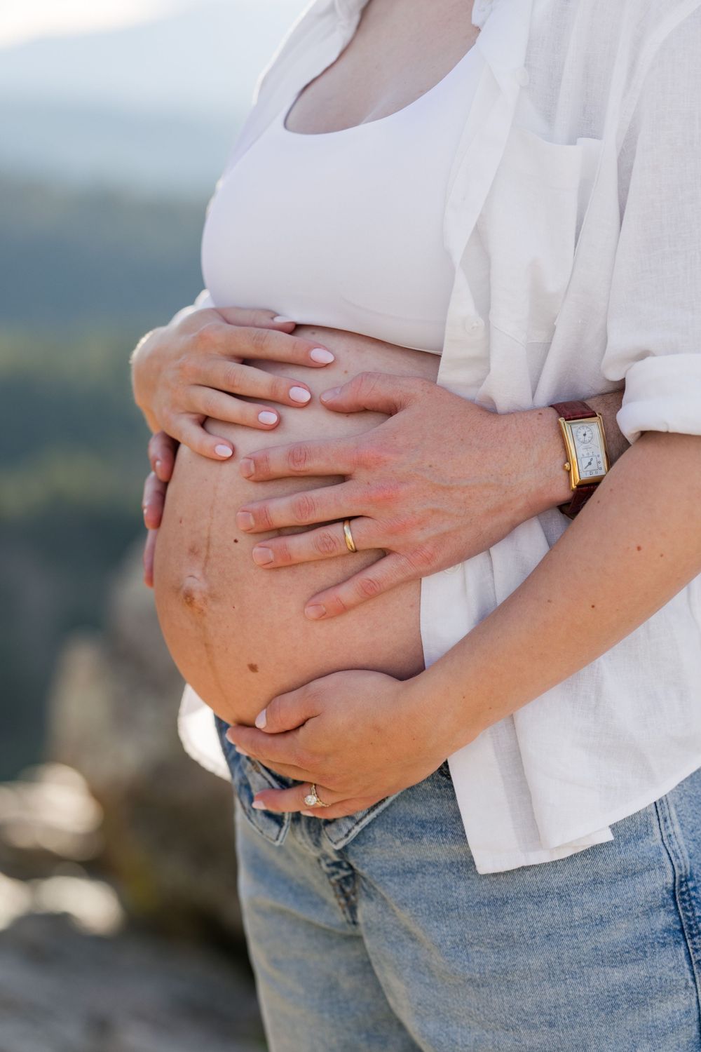 Pregnant woman in white shirt and jeans cradling baby bump outdoors with mountain backdrop.