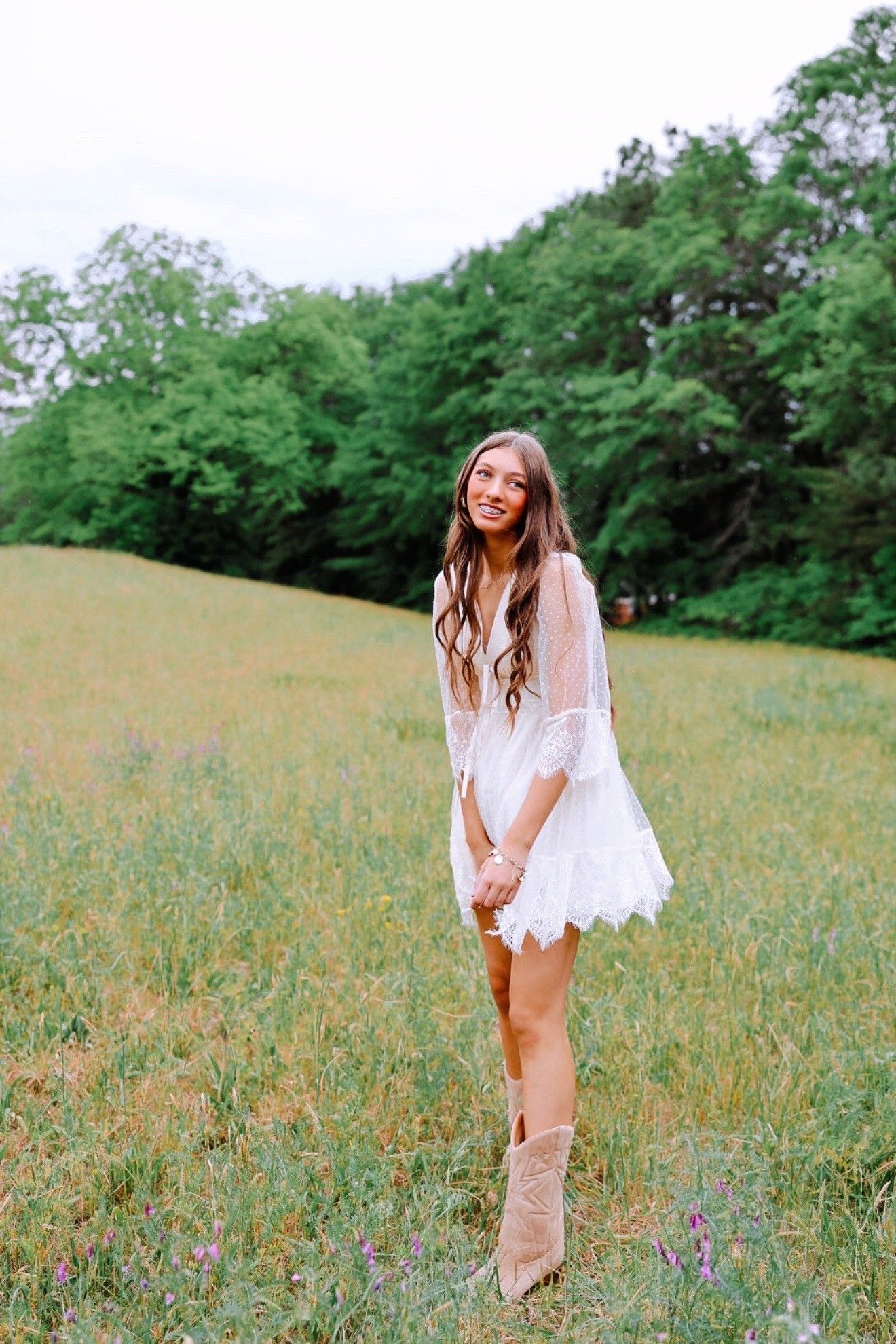 Smiling teenage girl with braces and long brown hair sitting in a grassy field wearing a floral dress and charm bracelet.