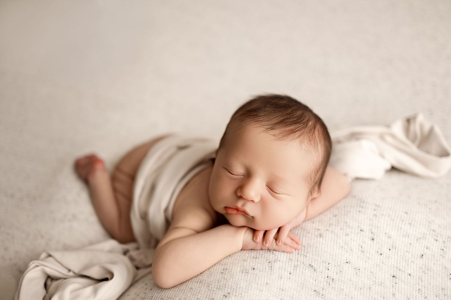 Peaceful newborn baby sleeping on cream knit blanket with hands tucked under chin in soft natural light photography.