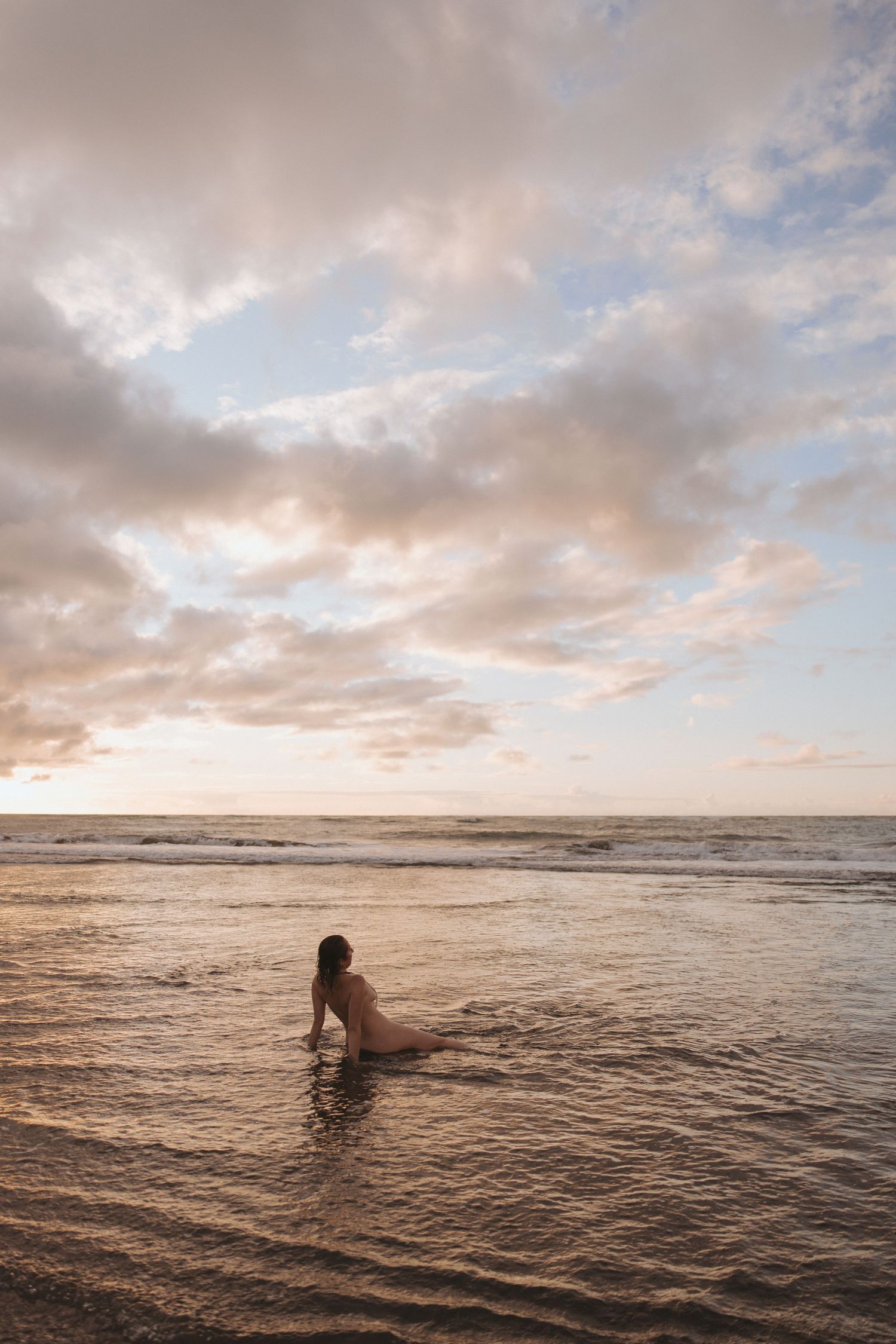 A woman sits in shallow ocean waves on a sandy beach during a golden sunset with dramatic cloudy sky.