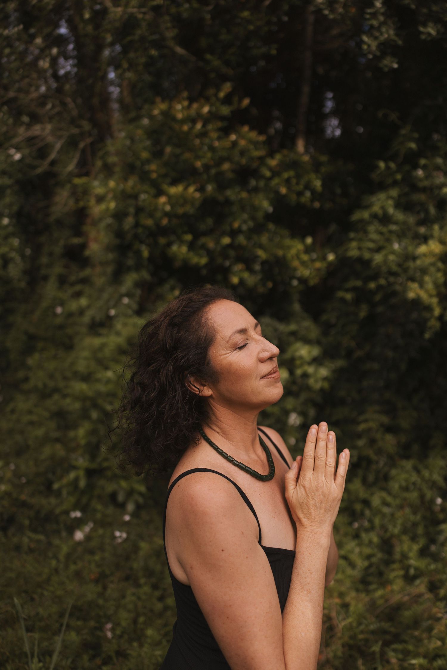 Woman meditating outdoors in prayer pose with eyes closed, surrounded by lush green trees in natural setting.