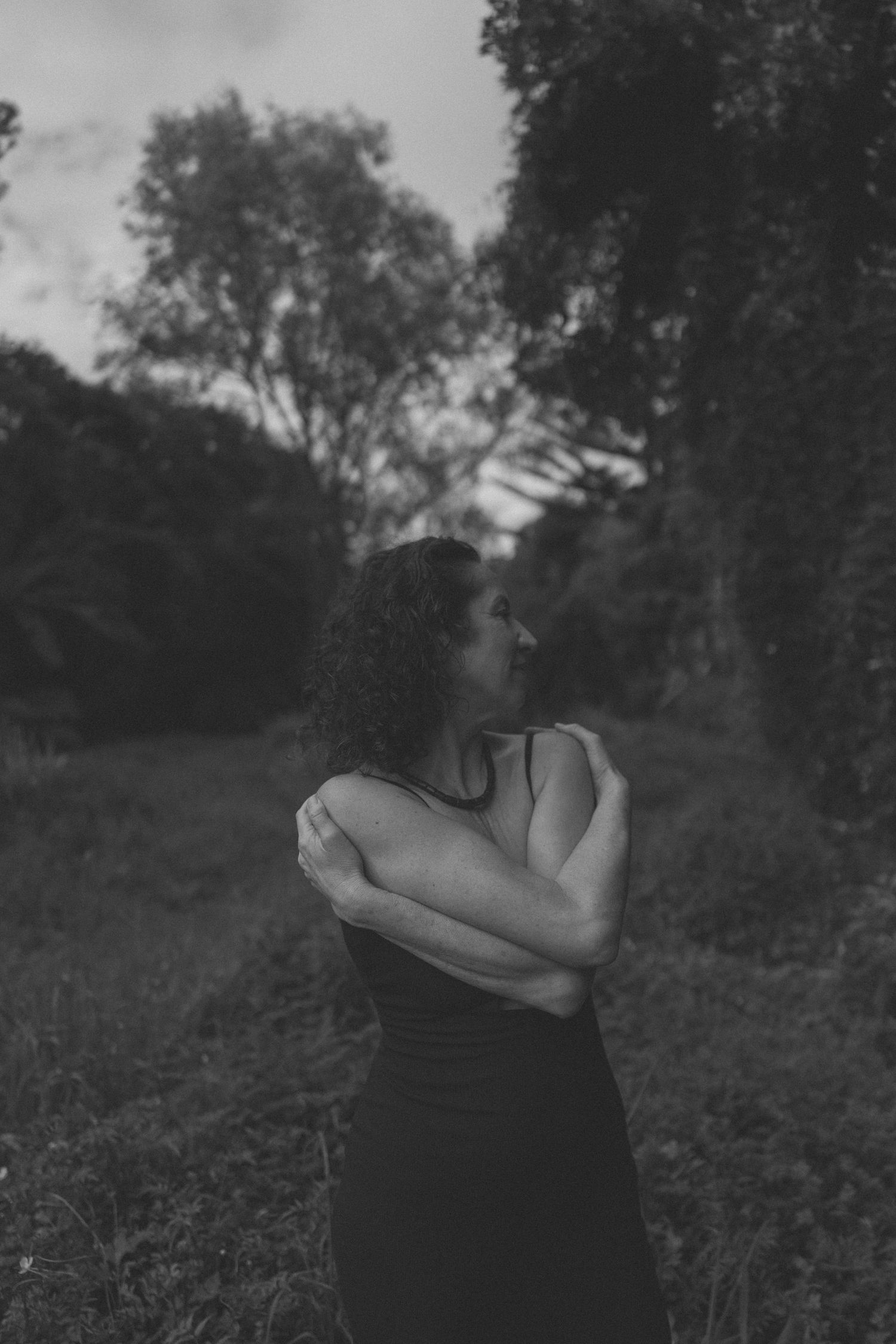 Black and white photo of a woman in a dark dress hugging herself in a moody outdoor forest setting.