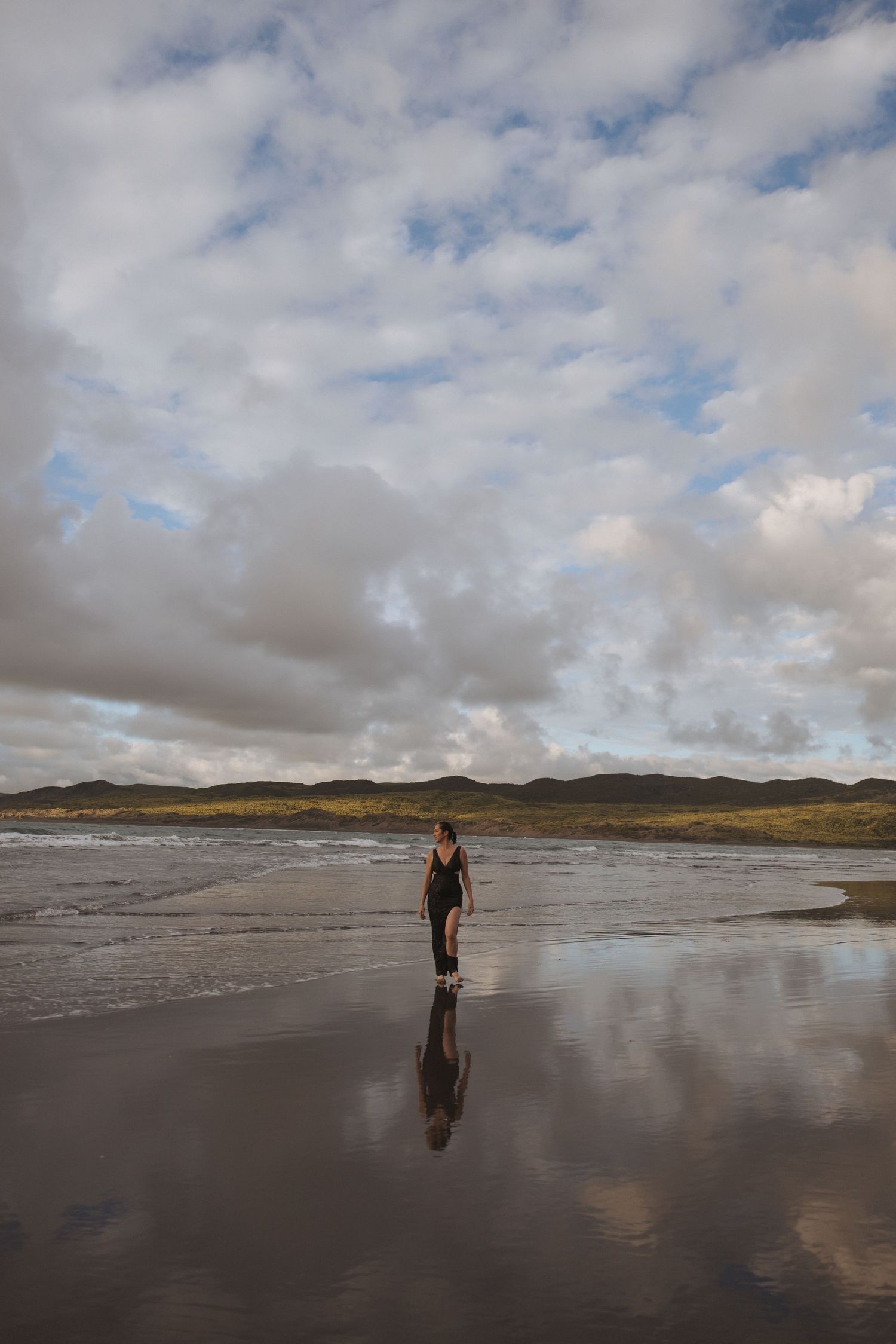 Woman in black swimsuit walks along a reflective wet beach with dramatic cloudy sky and green hills.