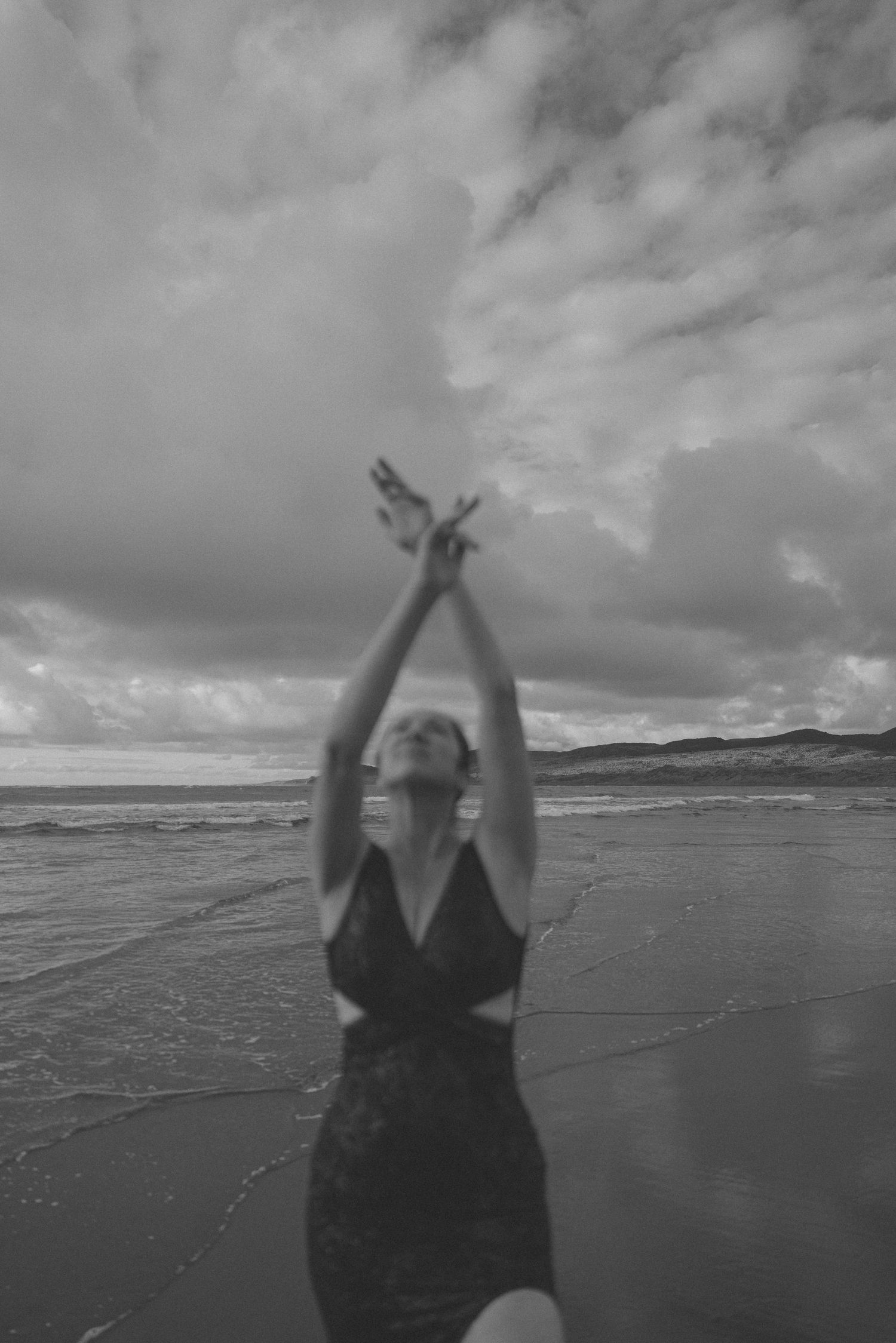 Black and white photo of woman in swimsuit raising arms toward dramatic cloudy beach sky.
