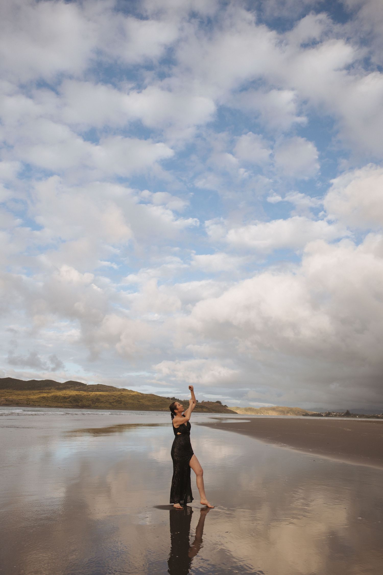 Woman in black dress dancing on reflective beach with dramatic cloudy sky and coastal landscape backdrop.