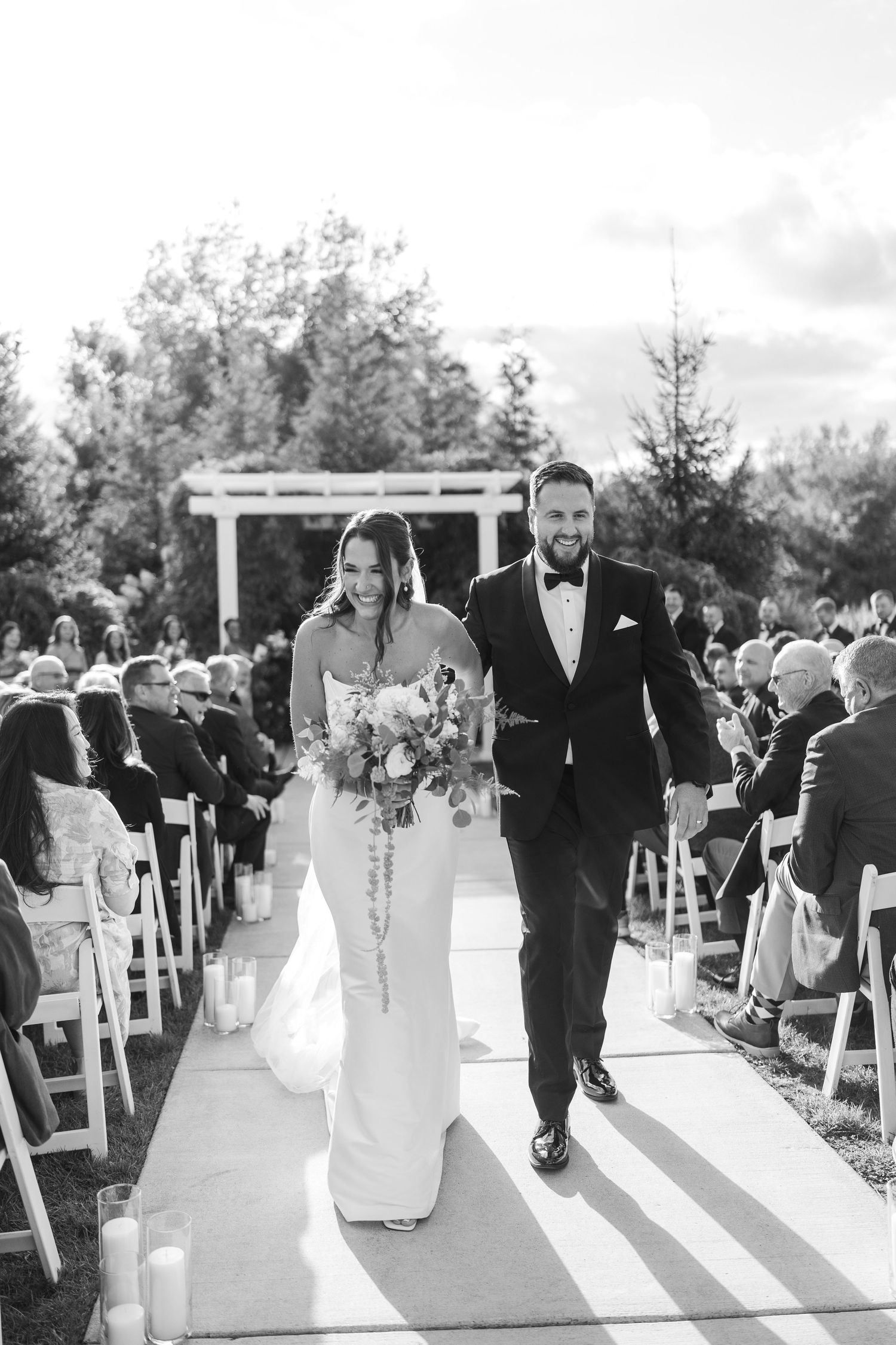 Outdoor wedding ceremony with couple at white arbor, guests seated in rows, surrounded by lush greenery and warm sunlight.