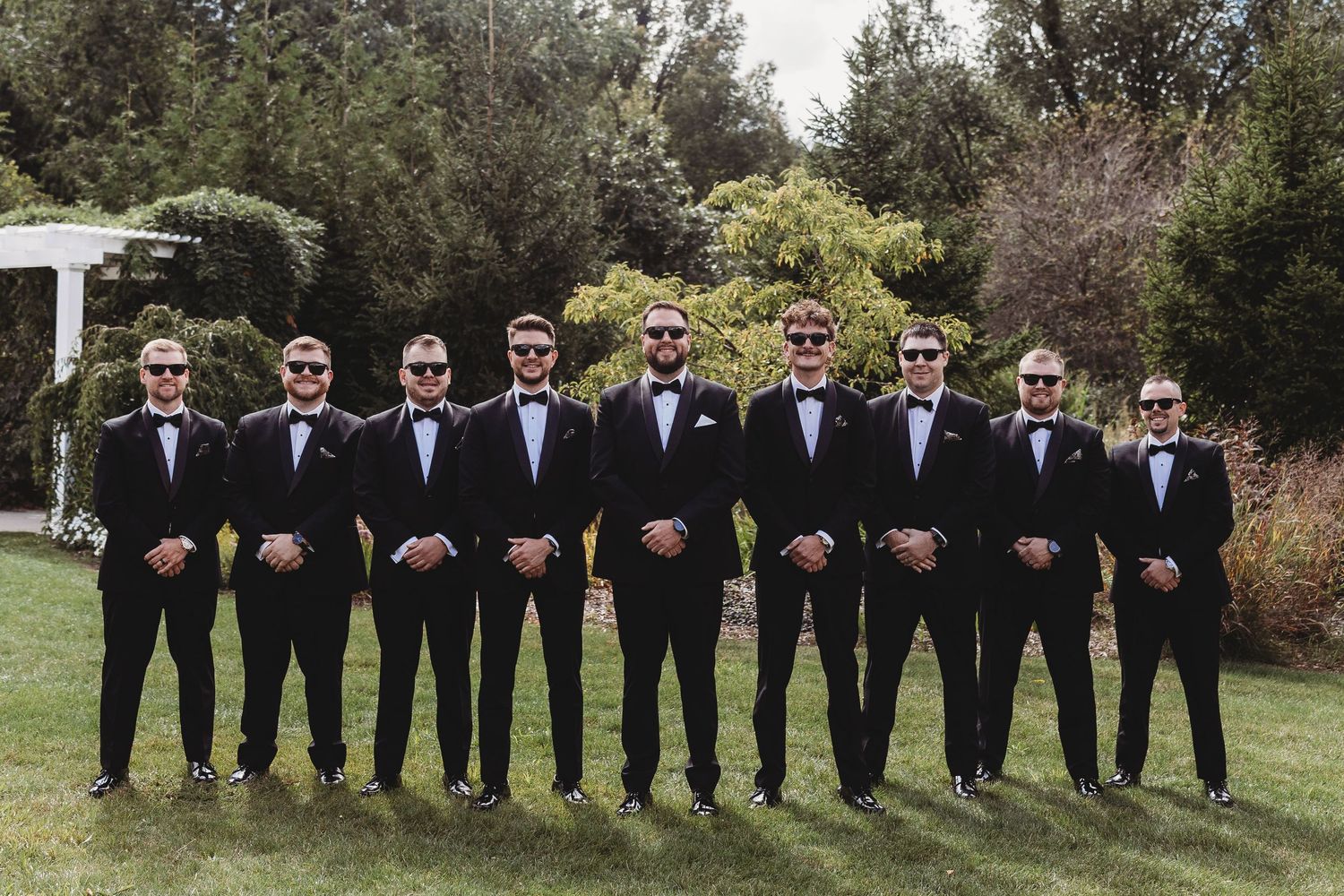 Groom and groomsmen in matching black tuxedos and sunglasses posing in a line outdoors.