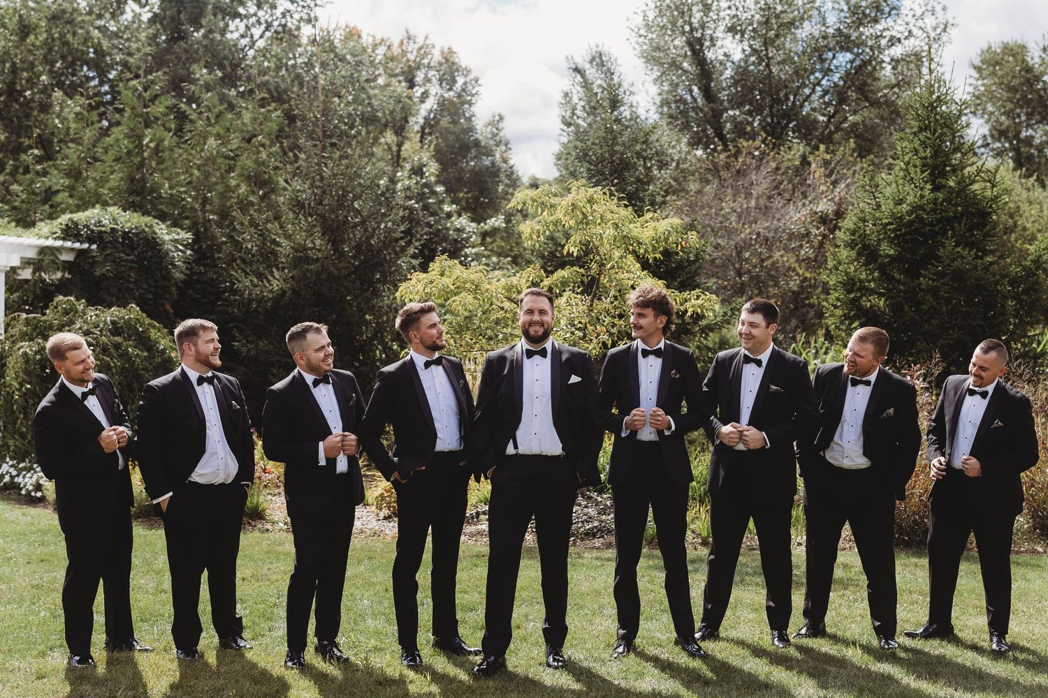Eight groomsmen in matching black tuxedos and bow ties laughing together outdoors on wedding day.