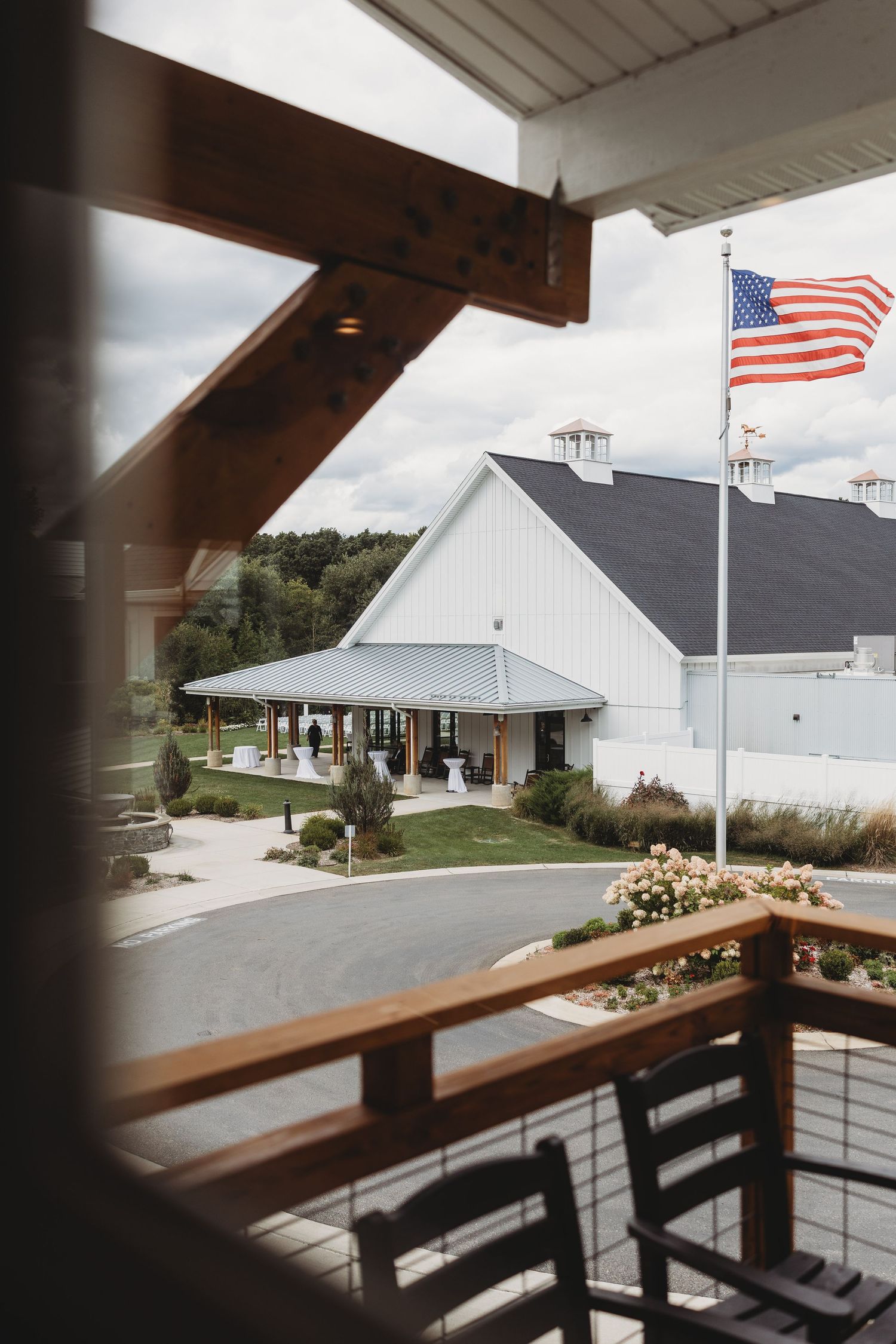 View from rustic wooden porch of white barn wedding venue with American flag flying on cloudy day.