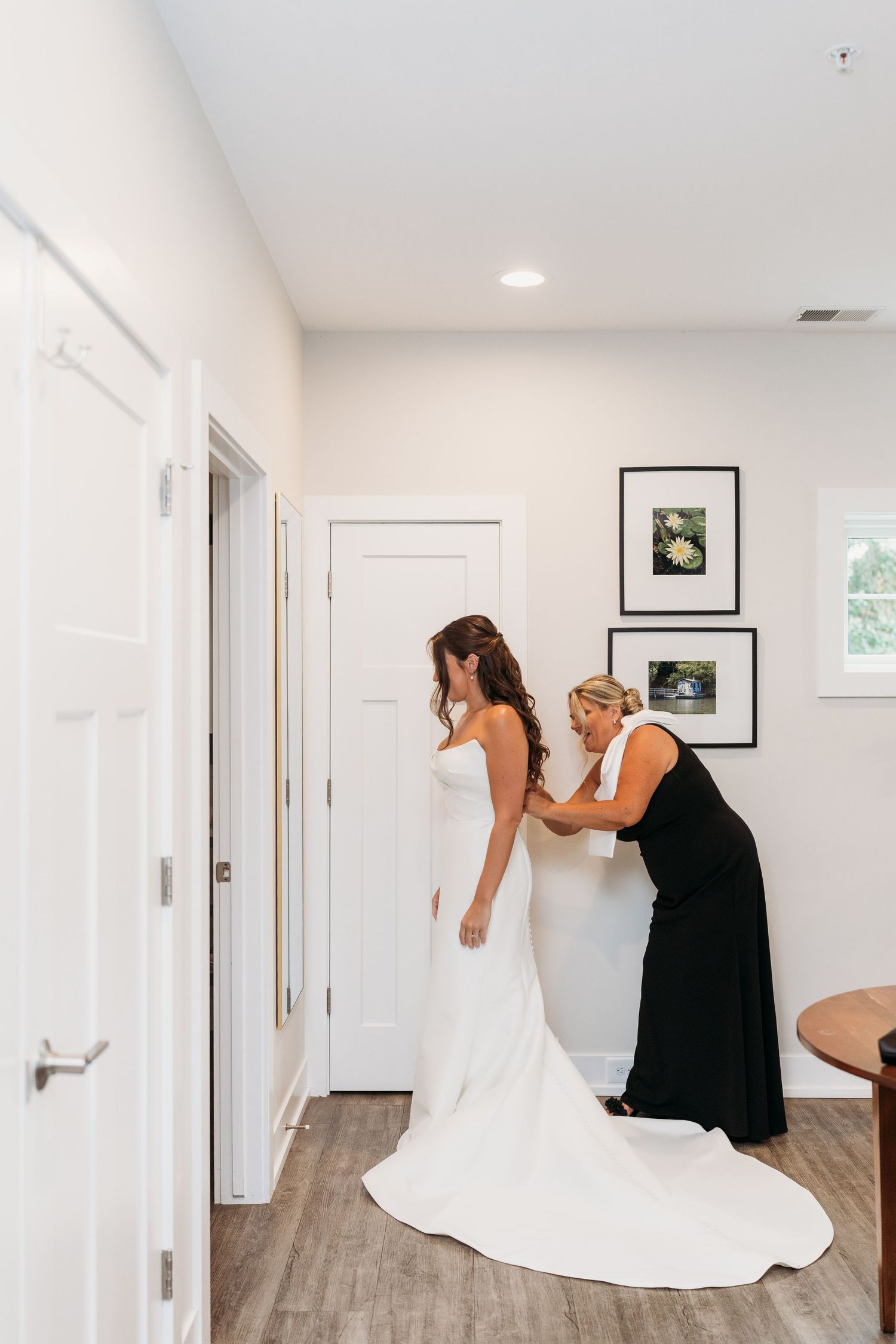 A woman in a black dress helps a bride zip up her elegant white wedding gown in a bright, modern bridal suite.