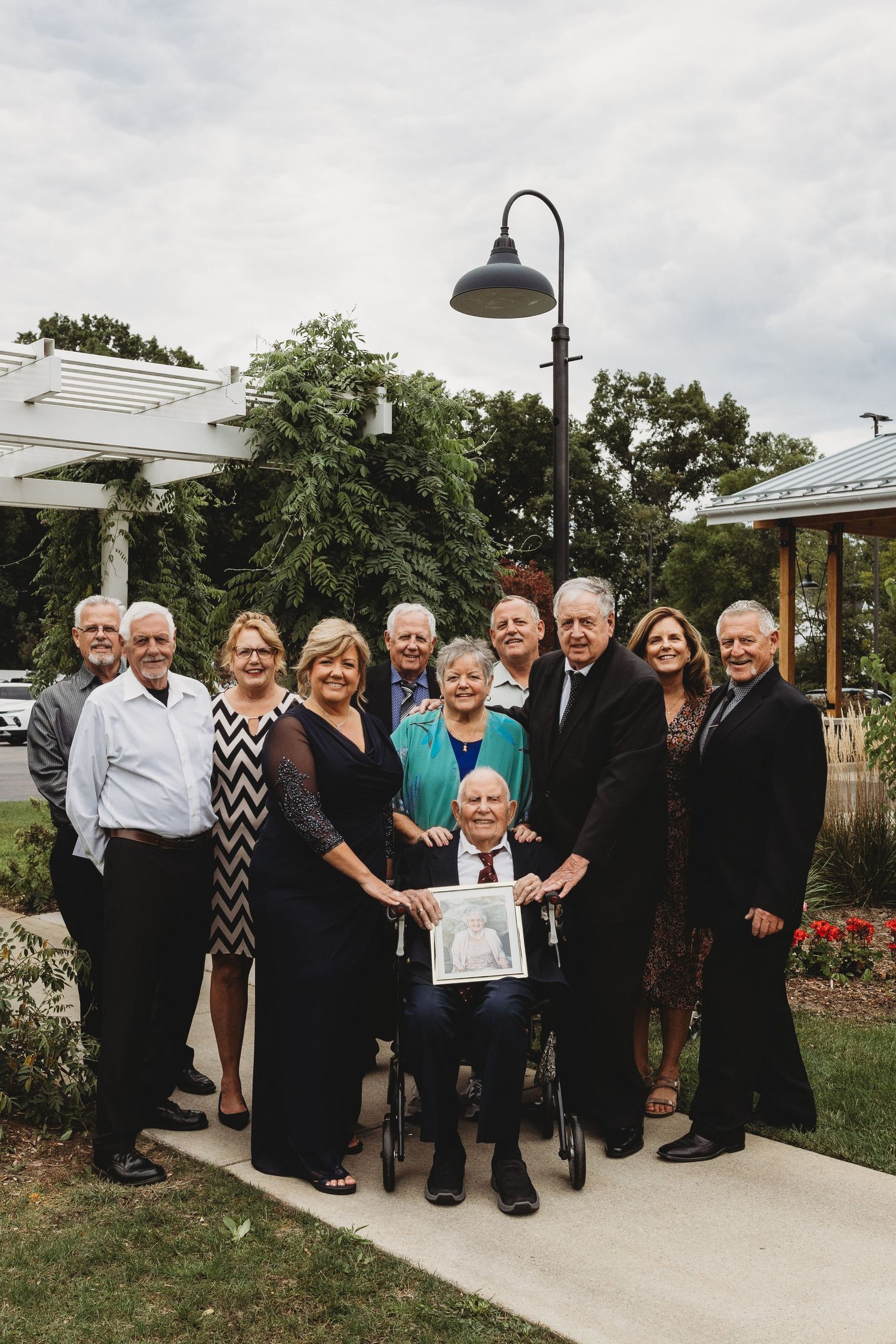 Family group portrait outdoors with elderly man in wheelchair holding framed photo, surrounded by relatives on a garden path.