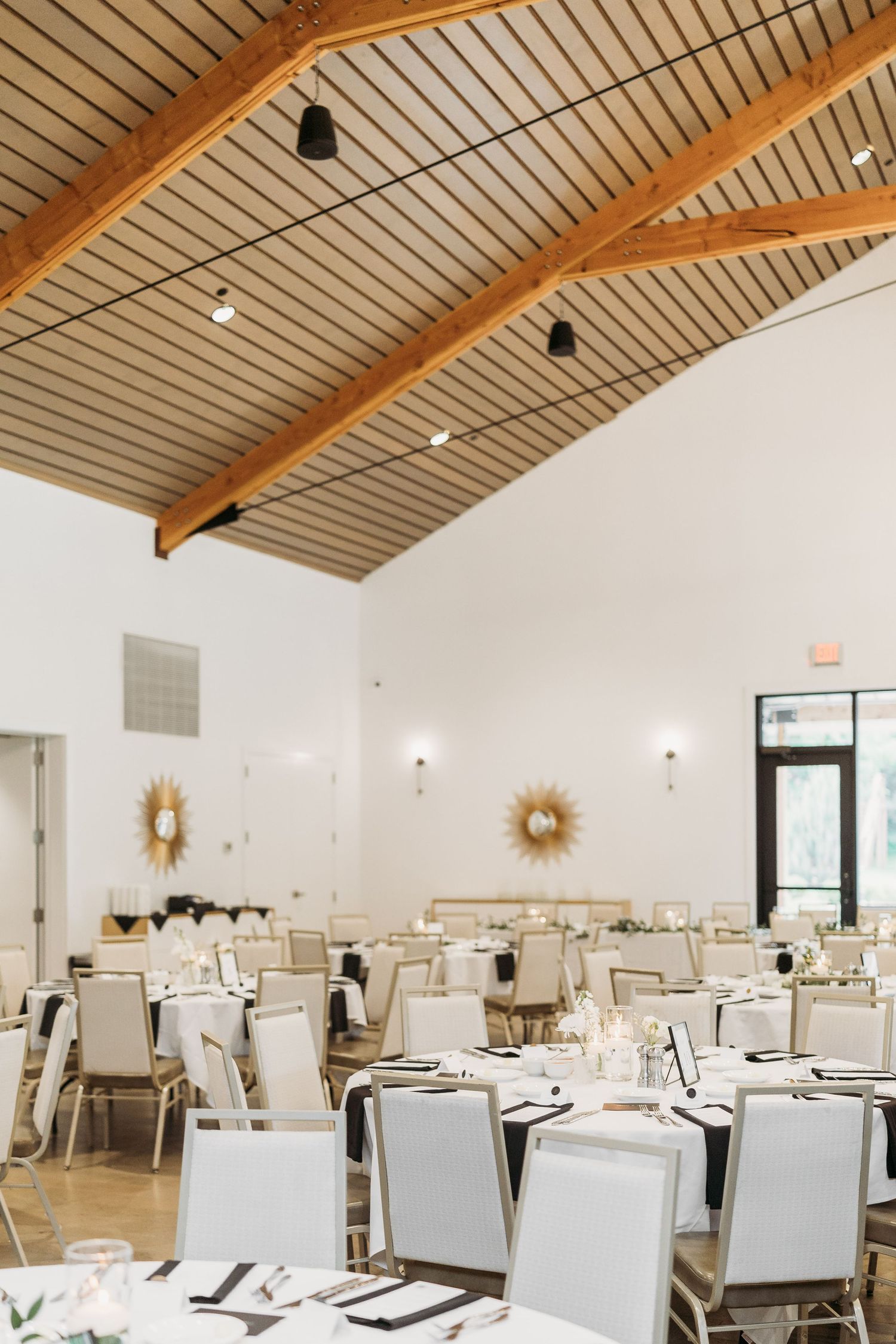 Bright wedding reception hall with vaulted wood beam ceiling, white chairs, and round decorated tables.