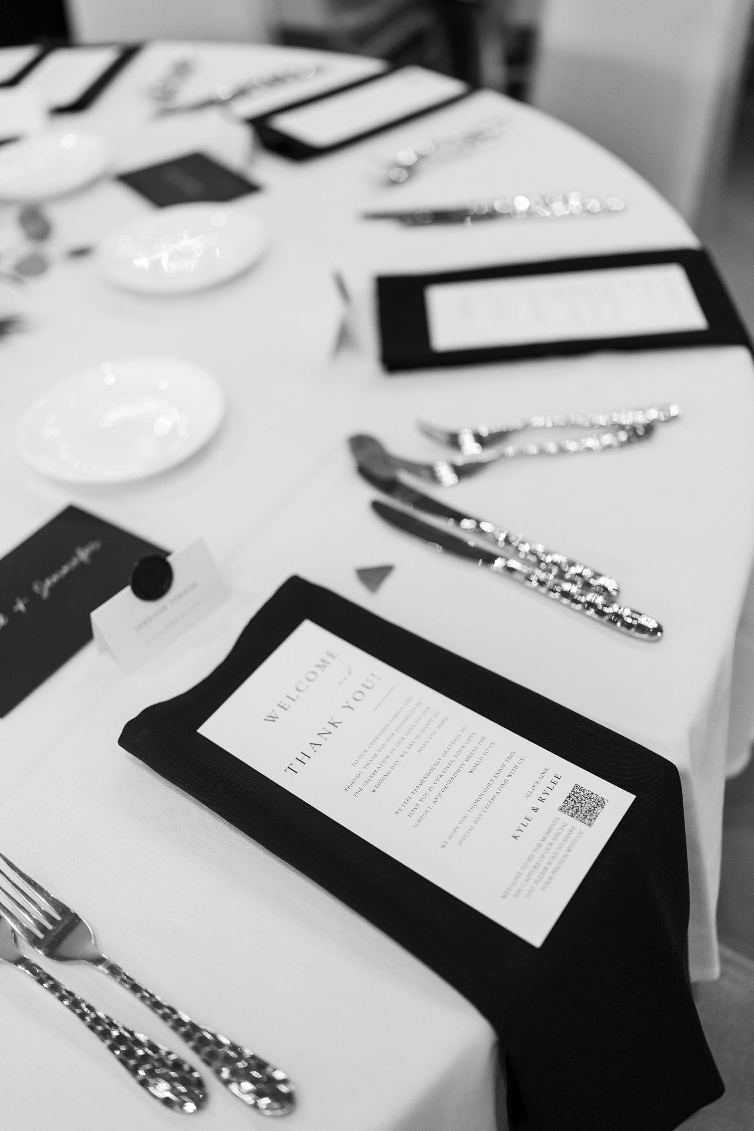 Elegant black and white wedding reception table setting with menus, silverware, and place cards on white linen.