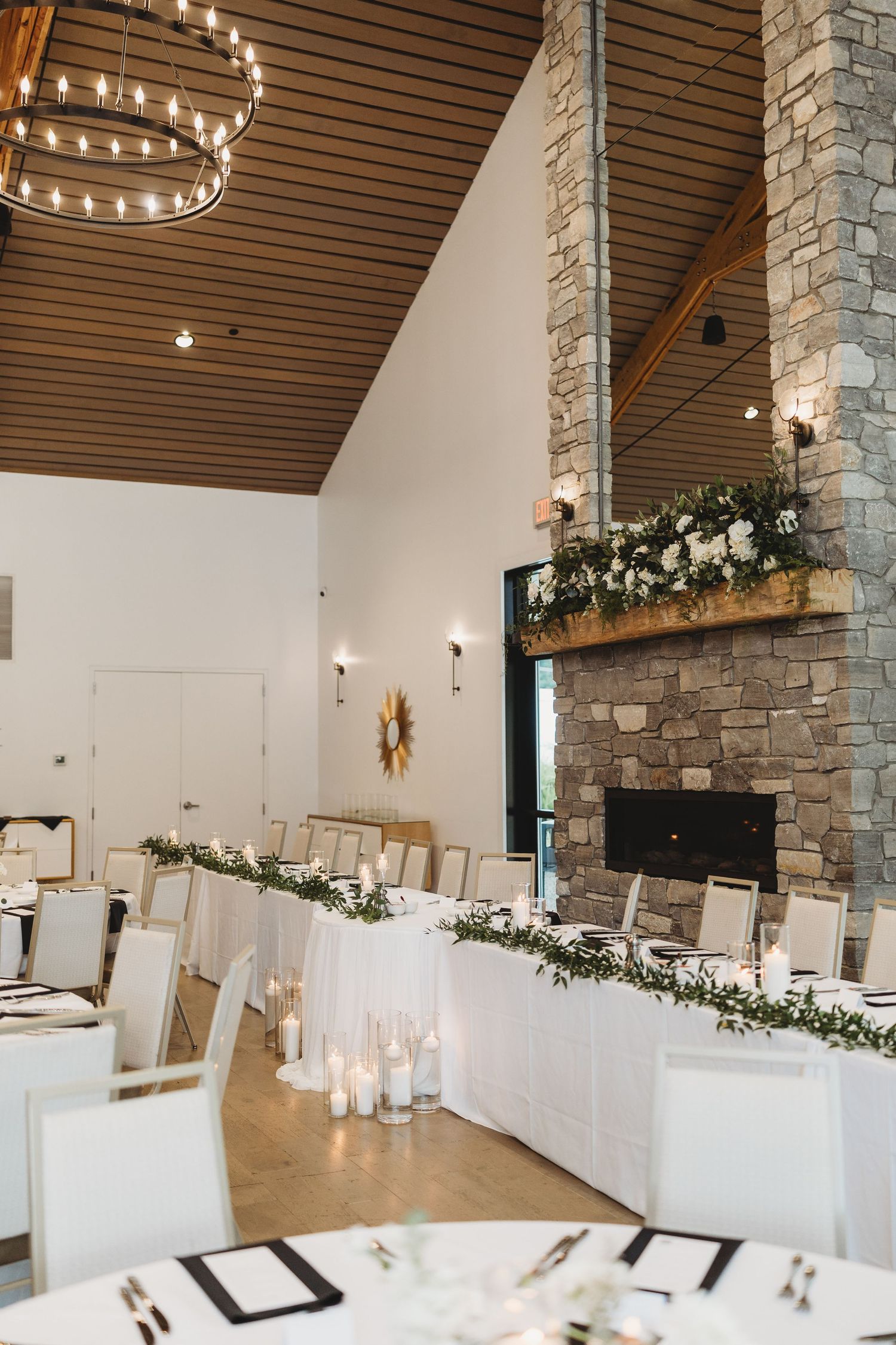 Elegant wedding reception hall with white tables, greenery garland, candles, and a stone fireplace adorned with floral arrangements.