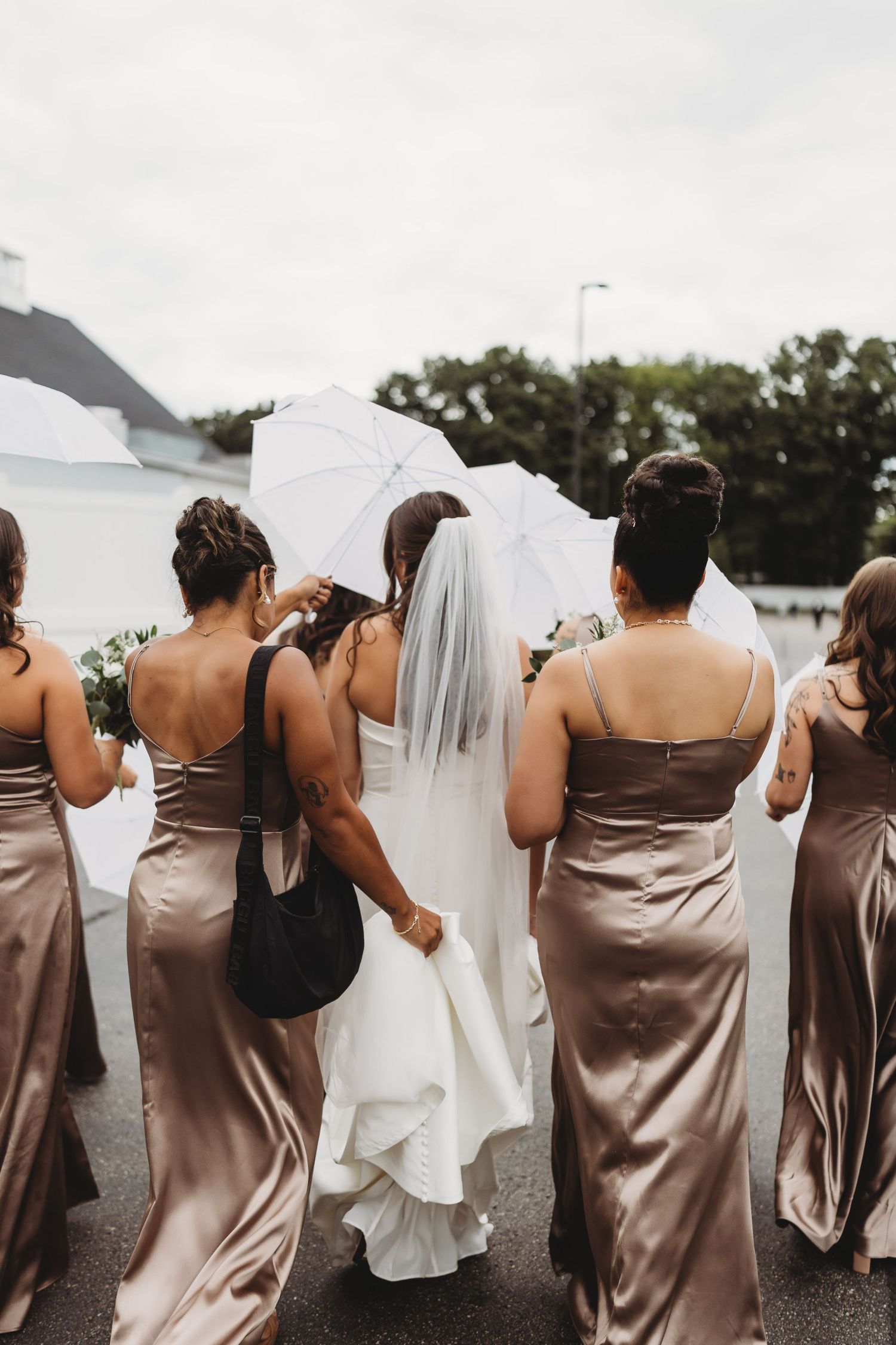 Bride in white gown with veil walks with bridesmaids in champagne satin dresses holding white parasol.