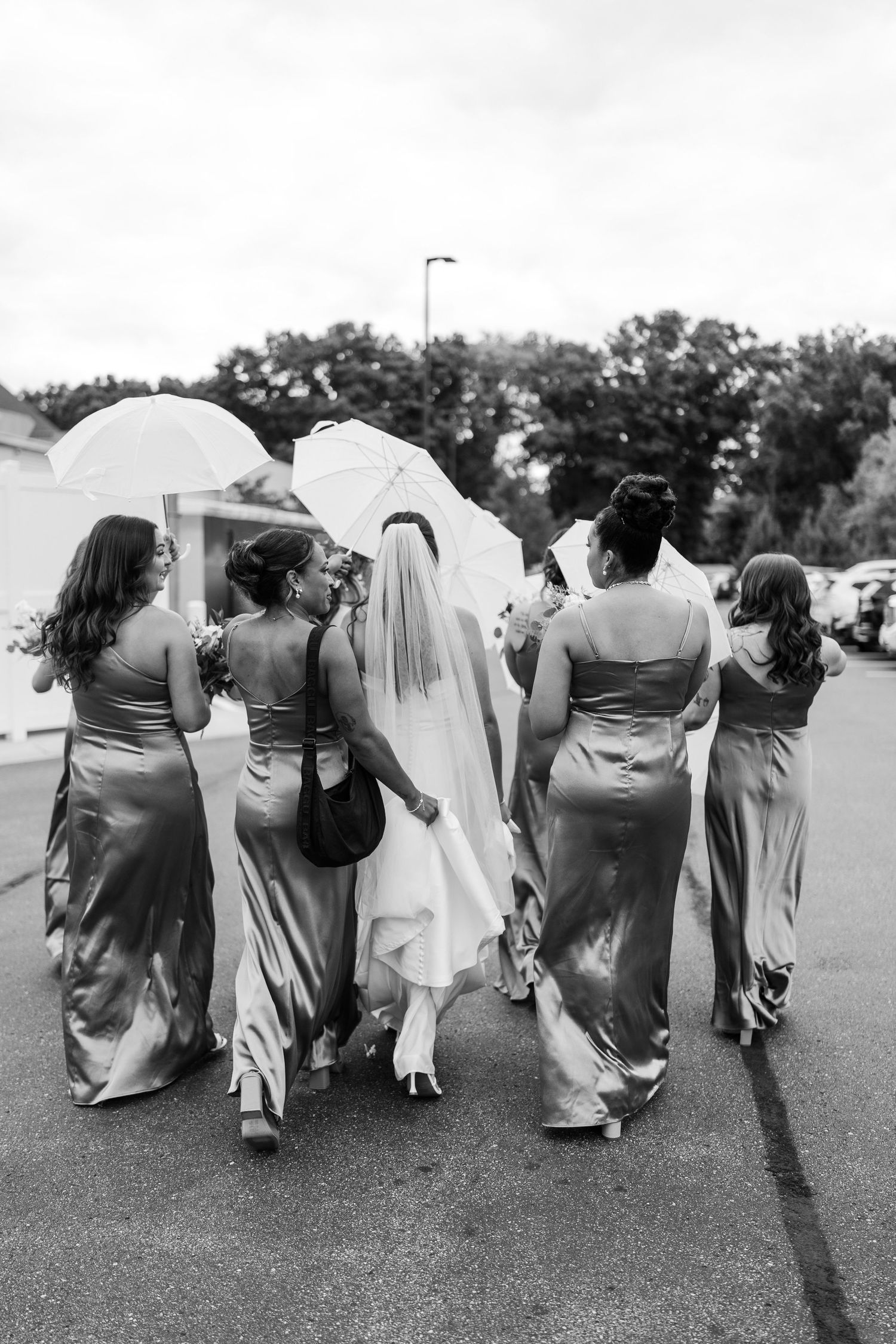 Black and white photo of bride and bridesmaids in satin gowns walking together, bride holding umbrella and veil flowing.