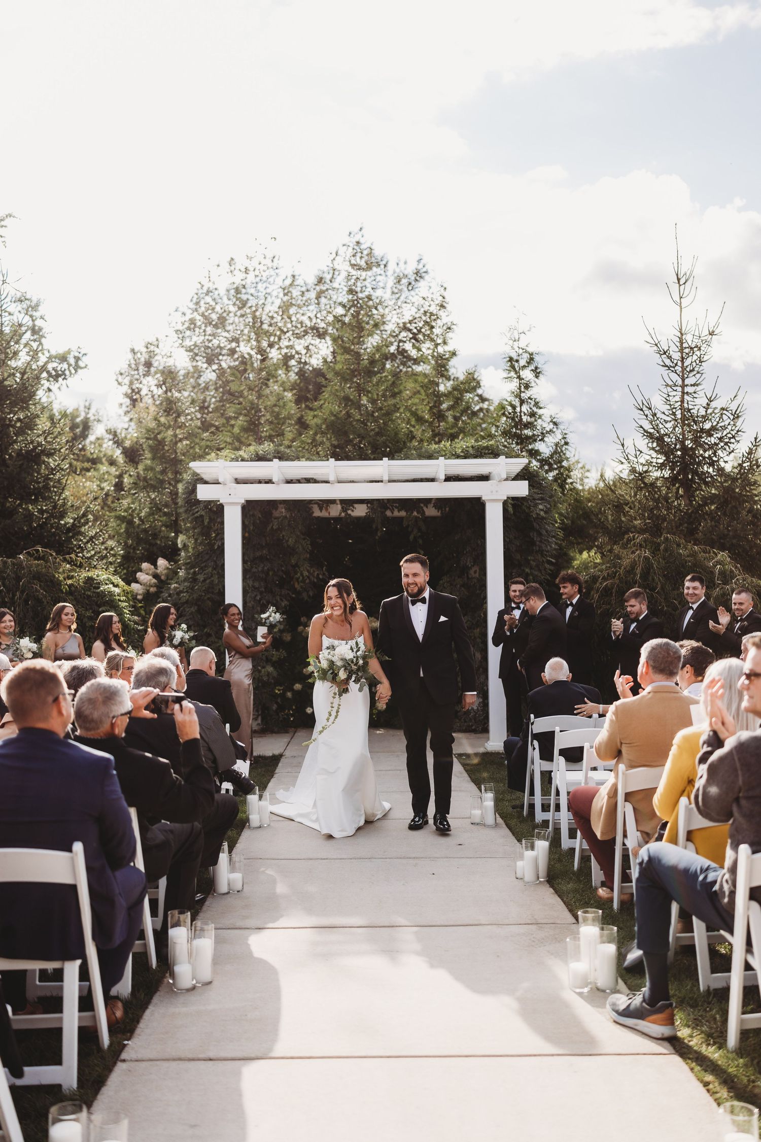 Bride and groom walk down outdoor wedding aisle lined with white chairs and candles beneath a white pergola.