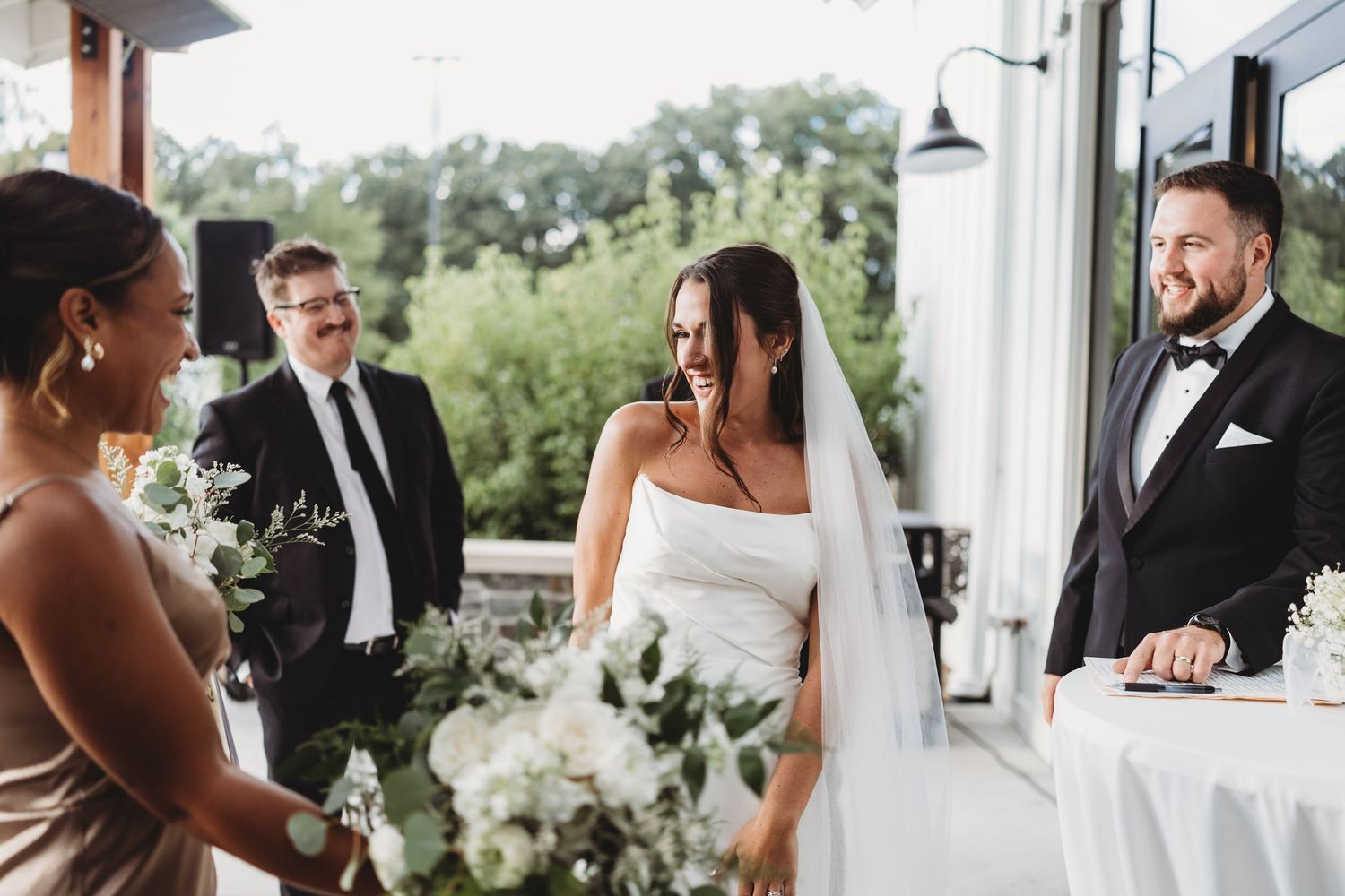 Bride laughing with groom and wedding party during indoor ceremony with floral bouquets and greenery backdrop.