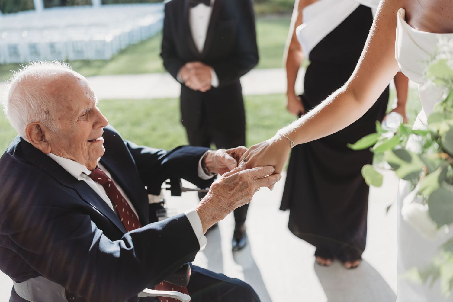 Elderly man in wheelchair holding hands with bride at outdoor wedding ceremony.