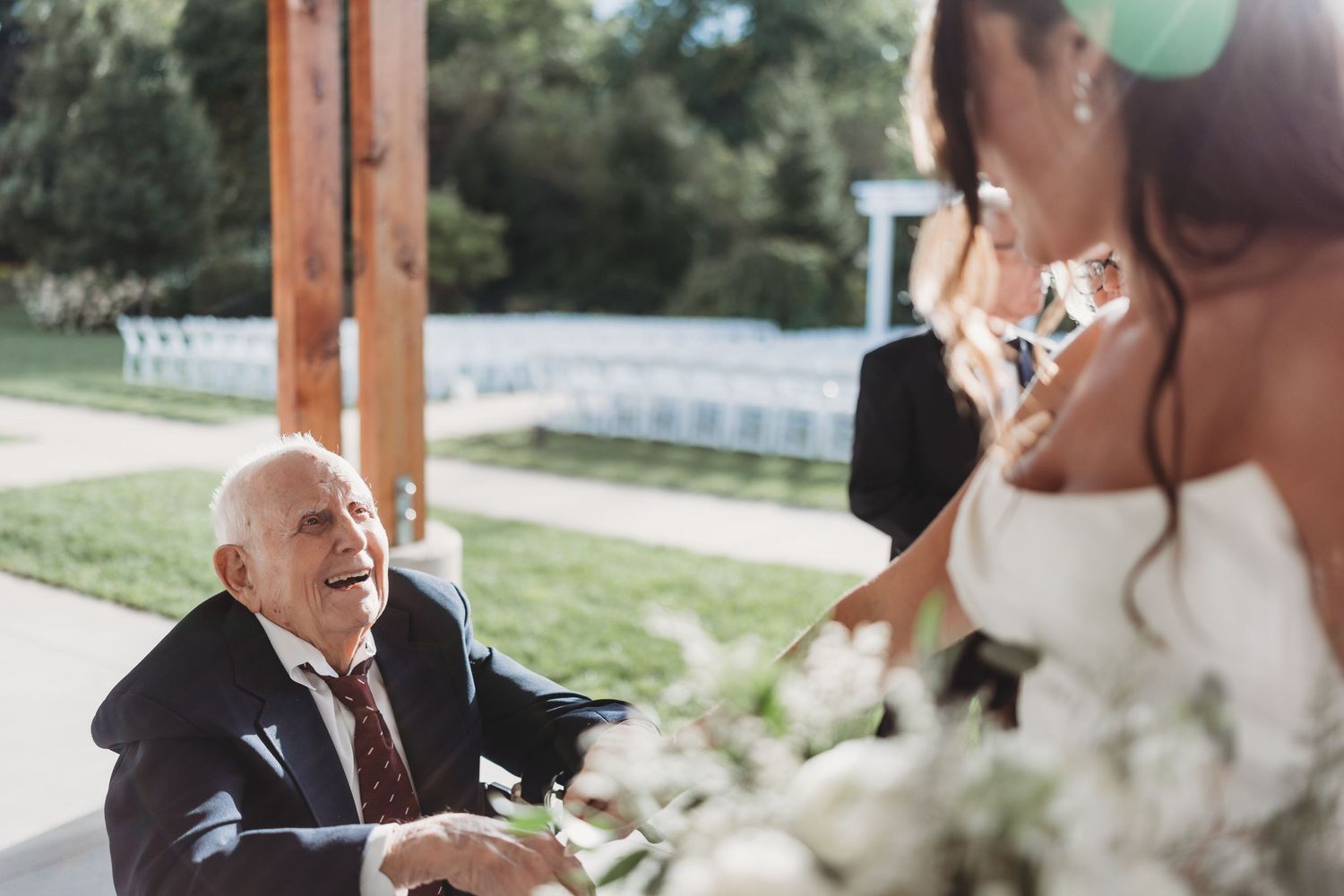 Elderly man in suit smiles joyfully at bride holding white flowers during outdoor wedding ceremony.
