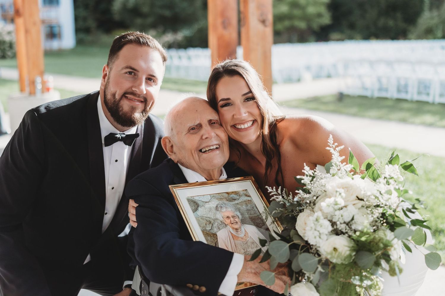 Bride and groom posing with elderly guest holding framed photo at outdoor wedding ceremony.