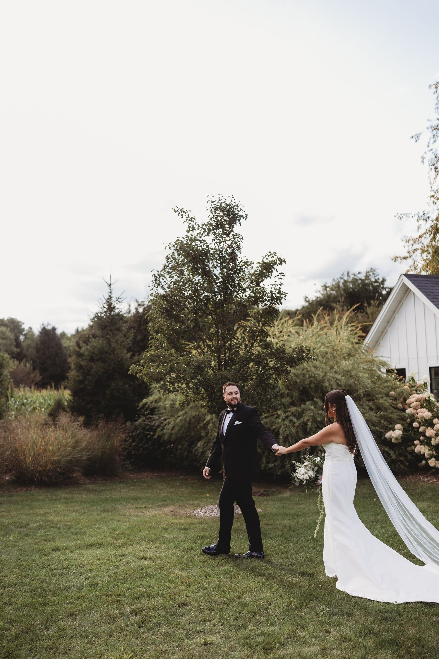 Bride and groom share a joyful moment on green lawn with white barn and lush trees in background.