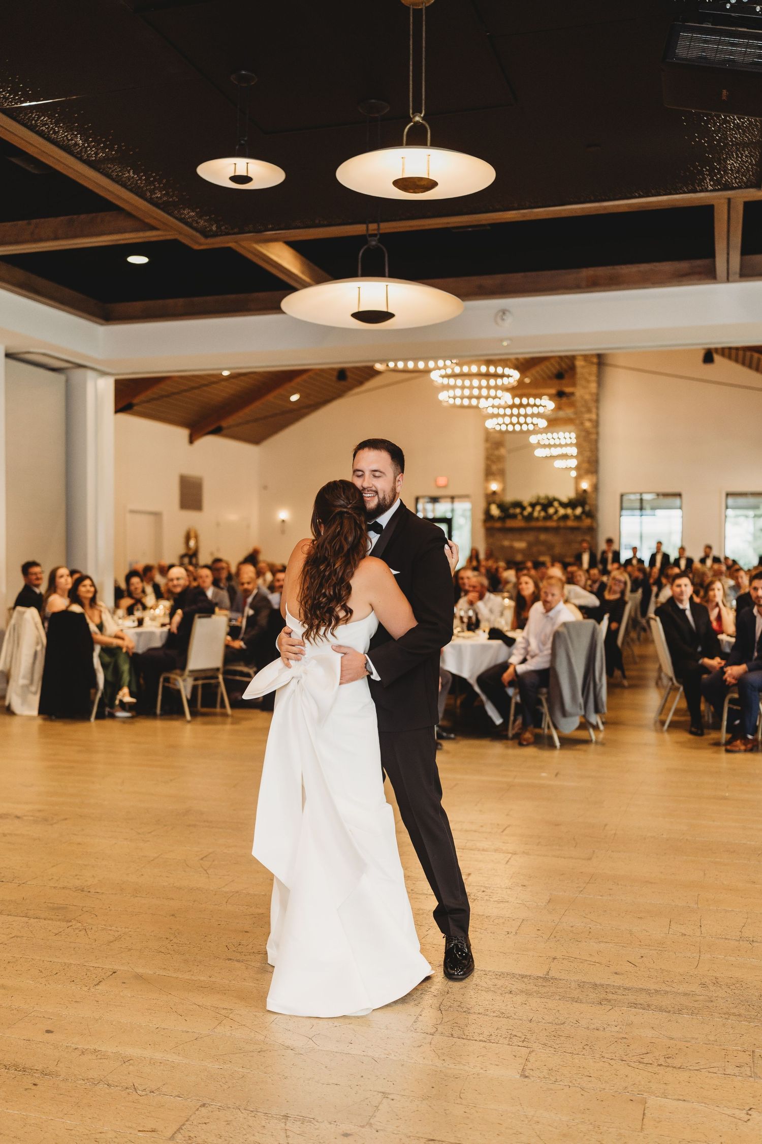 Bride and groom share their first dance on a hardwood floor surrounded by seated wedding guests in a elegant venue.