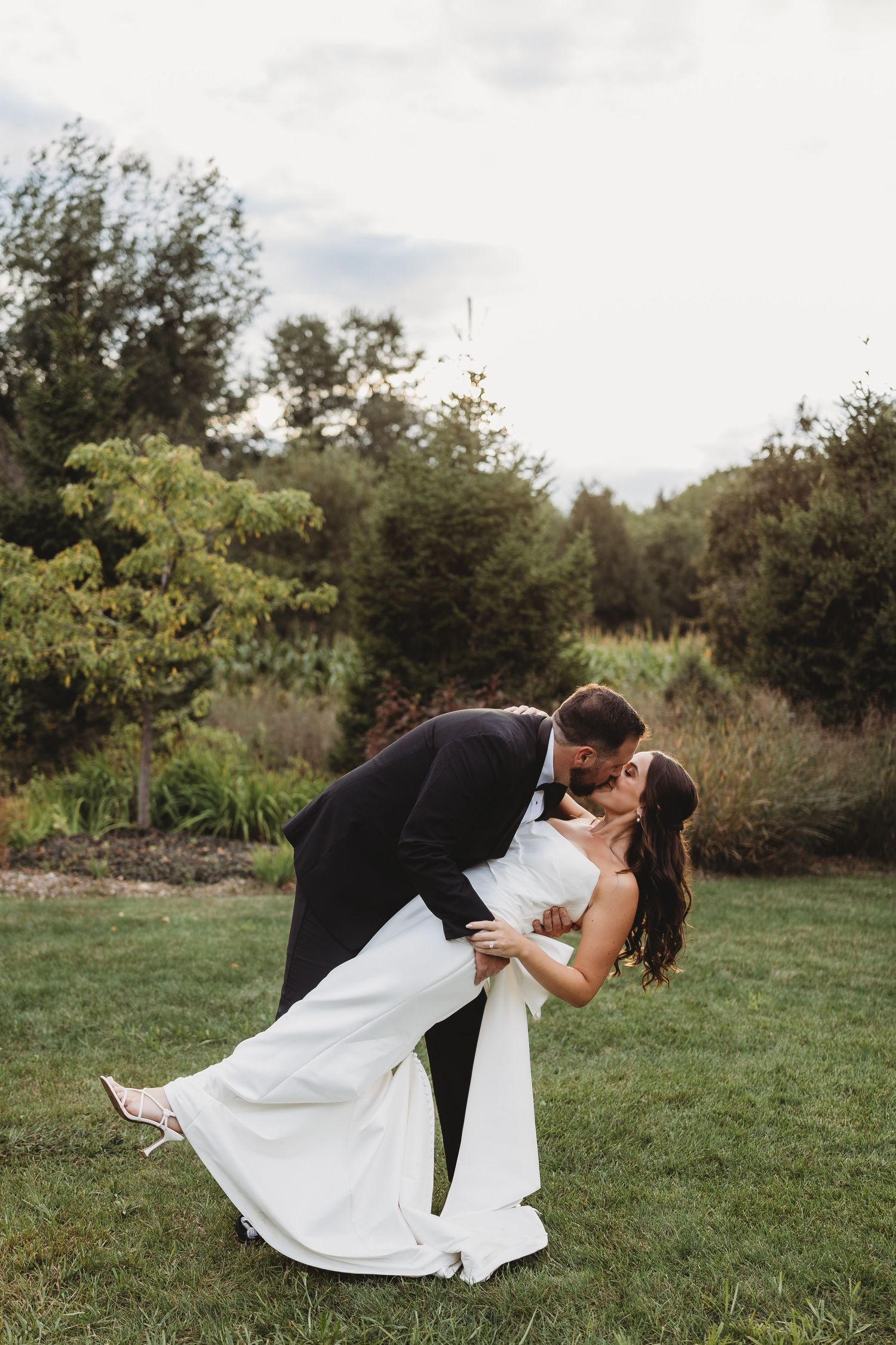 A groom in a black tuxedo dips his bride in a white gown for a romantic kiss on a lush green lawn.
