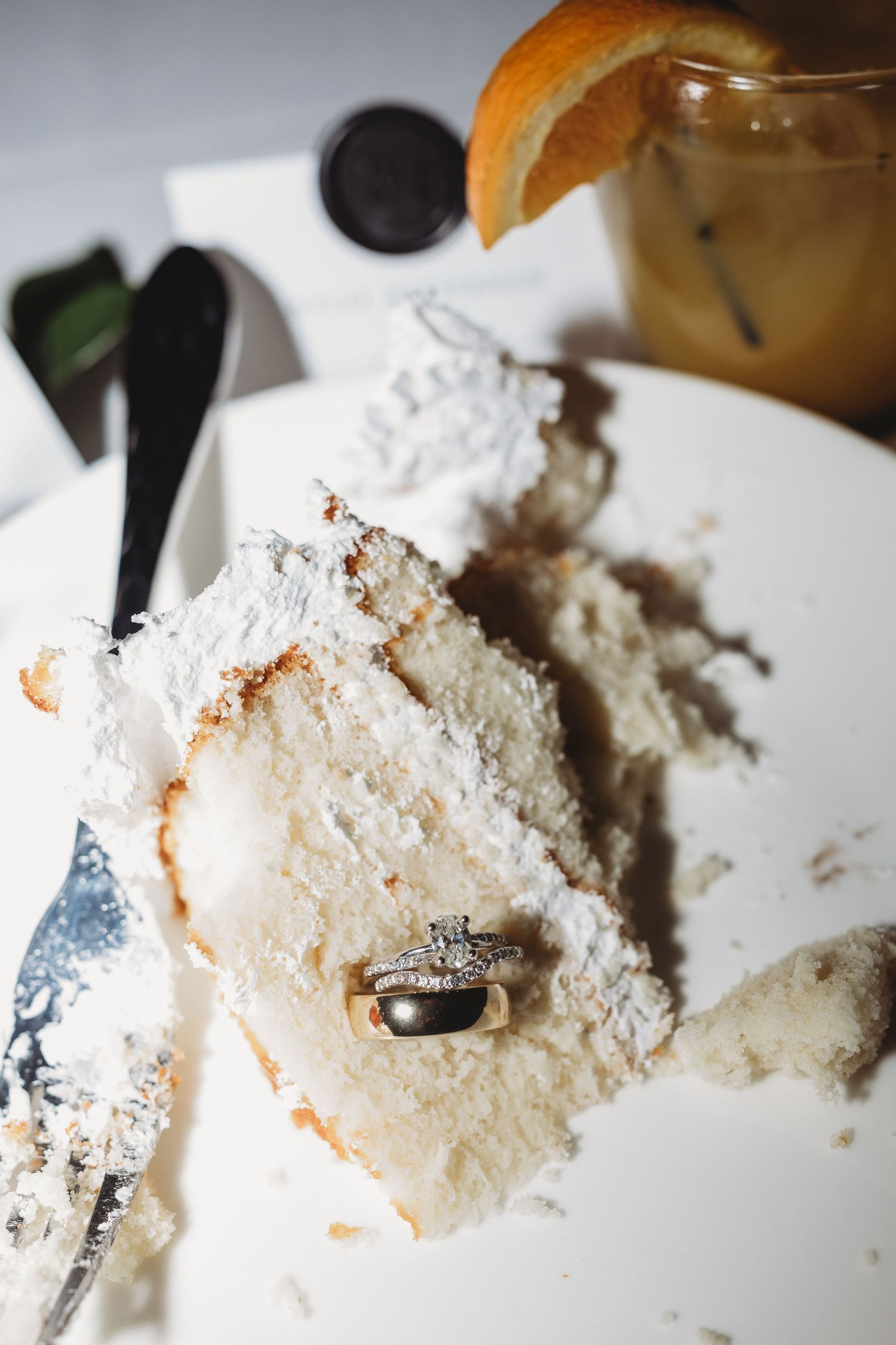 Wedding rings resting on slice of white coconut cake beside cocktail with orange garnish.