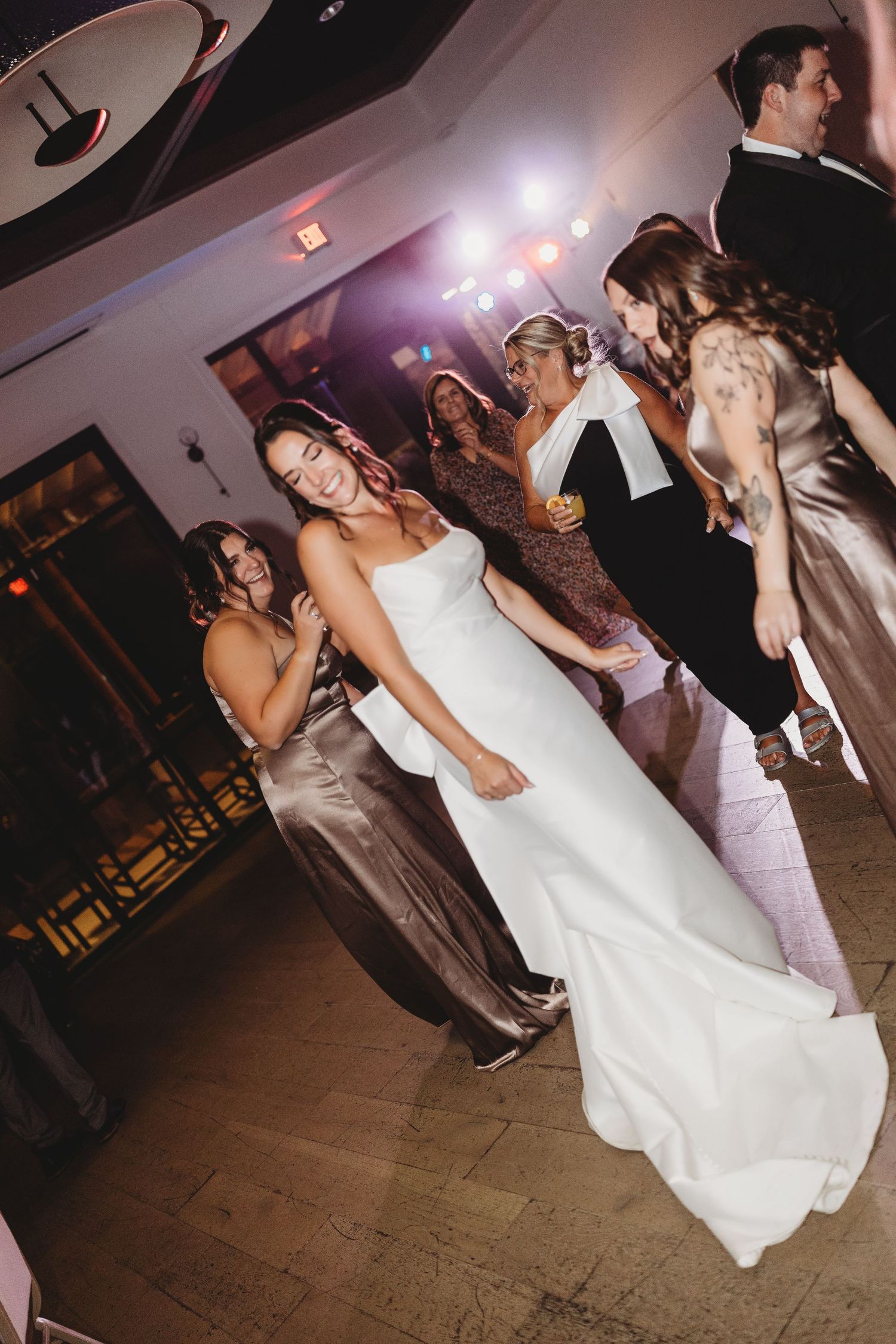 Bride in white gown dancing joyfully with guests on lit wedding reception dance floor.