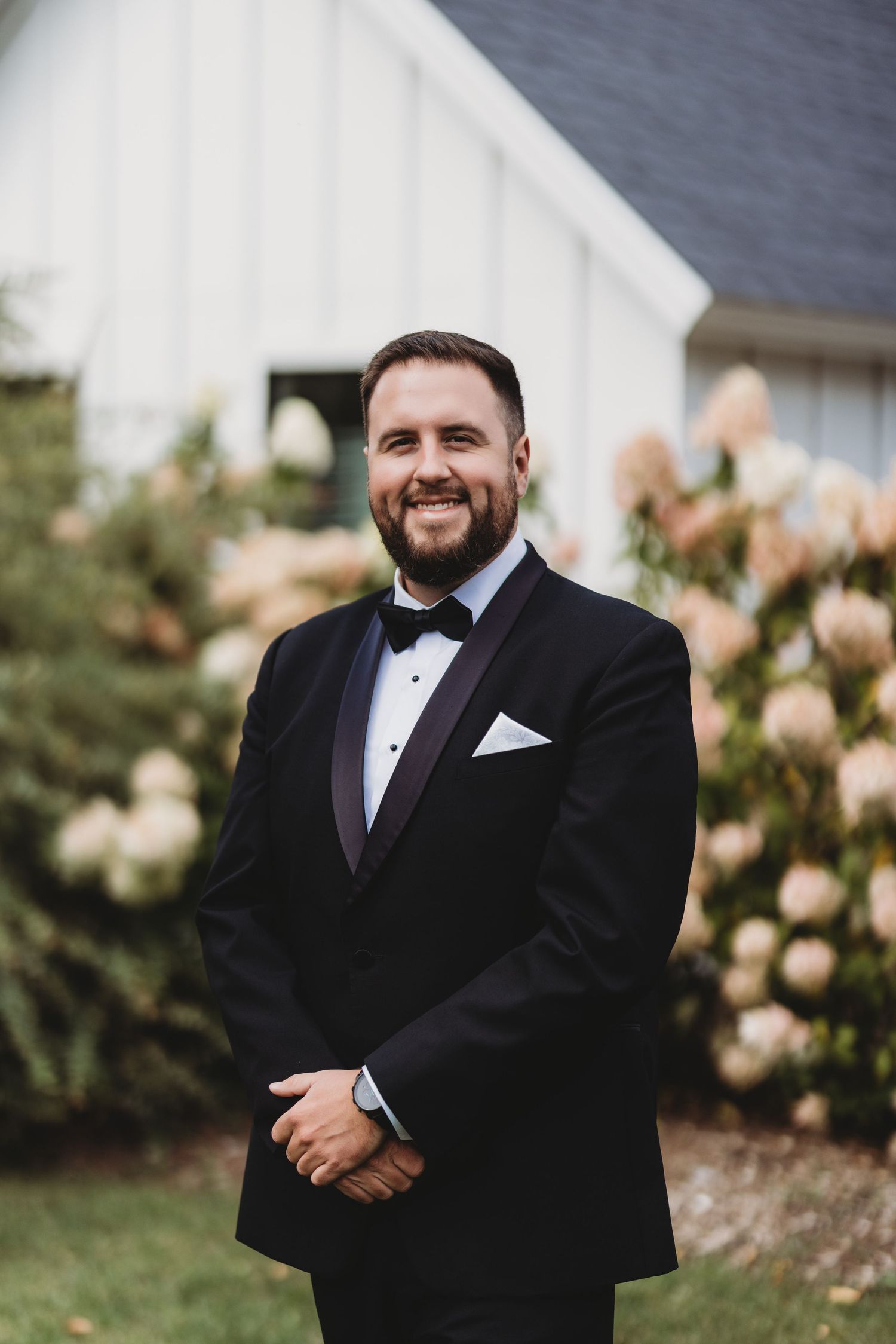 Groom in black tuxedo smiling in front of white barn and hydrangea bushes.