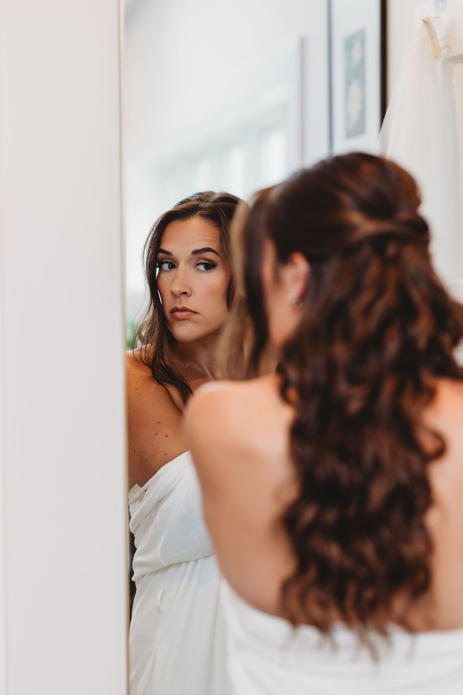 Bride with long brunette hair examining her reflection in a mirror while getting ready.