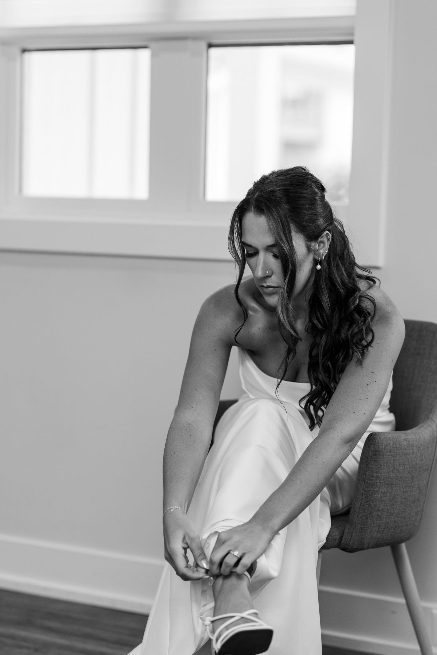 Bride in white gown puts on shoes while seated in black and white photo.