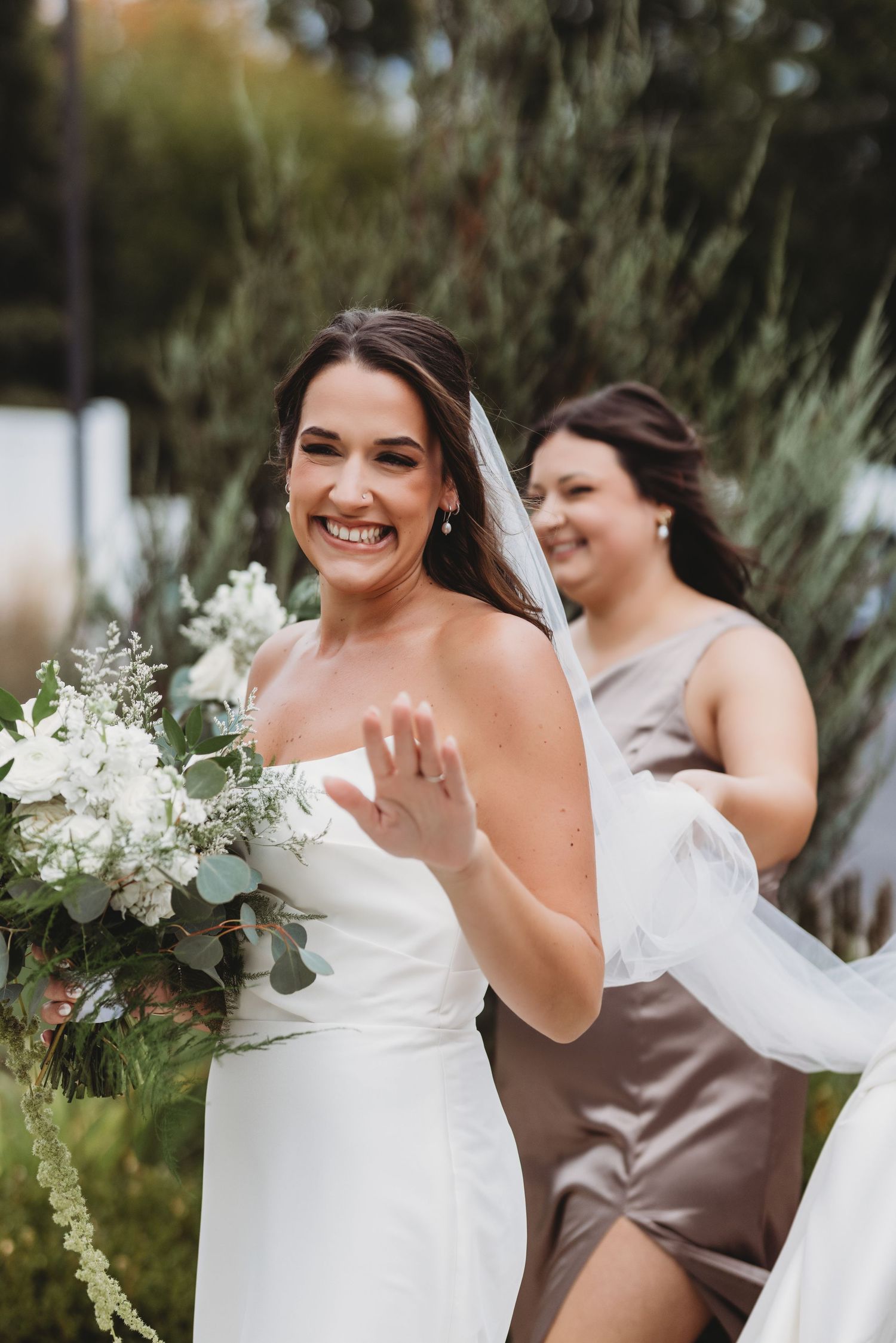 Smiling bride in strapless gown holding white floral bouquet with bridesmaid adjusting veil outdoors.