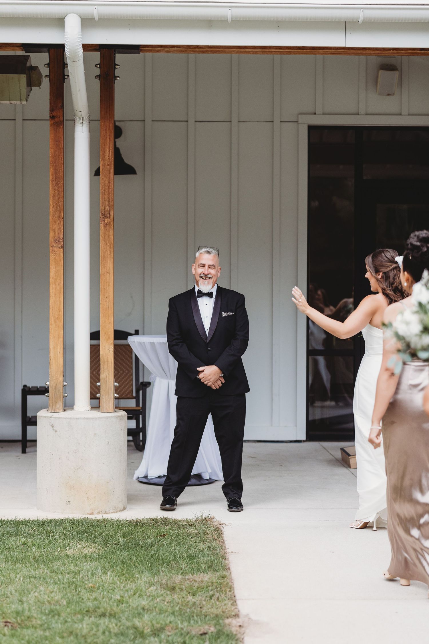 Smiling father in tuxedo awaits bride outside white barn venue during wedding day preparations.