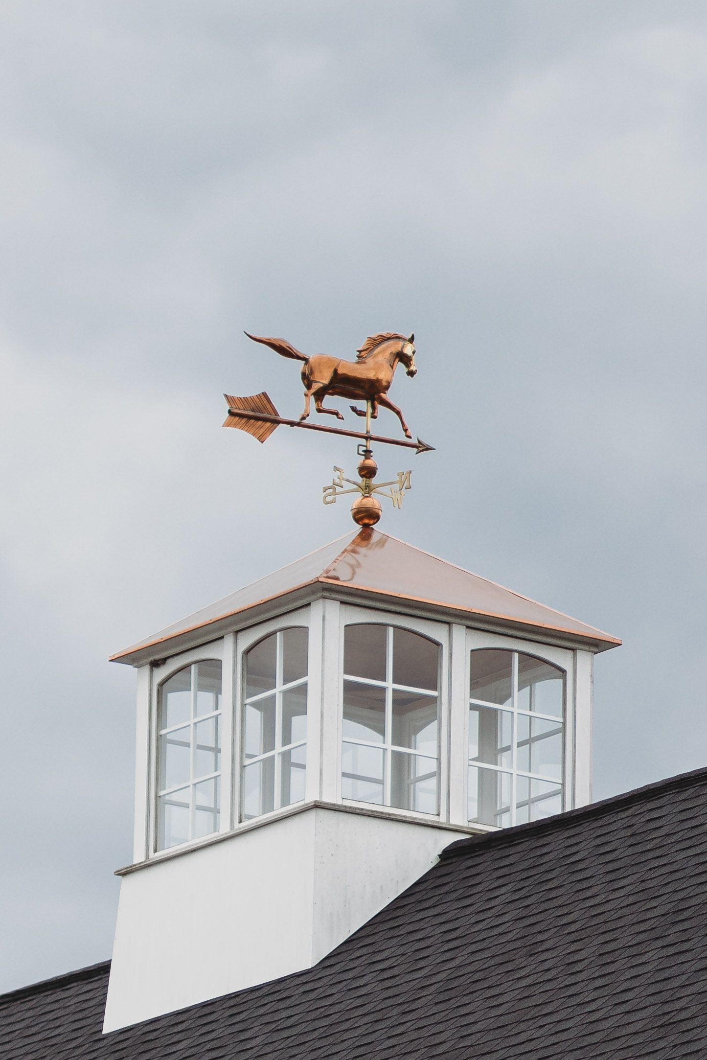 Copper horse weather vane atop a white cupola with windows on a dark shingled roof against cloudy sky.