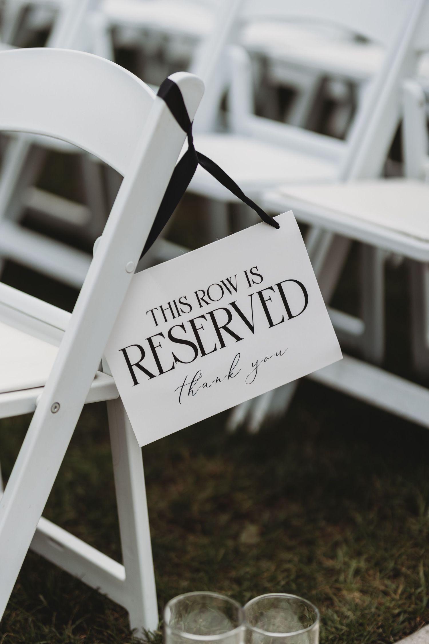 White folding chair with elegant reserved row sign tied with black ribbon at an outdoor wedding ceremony.