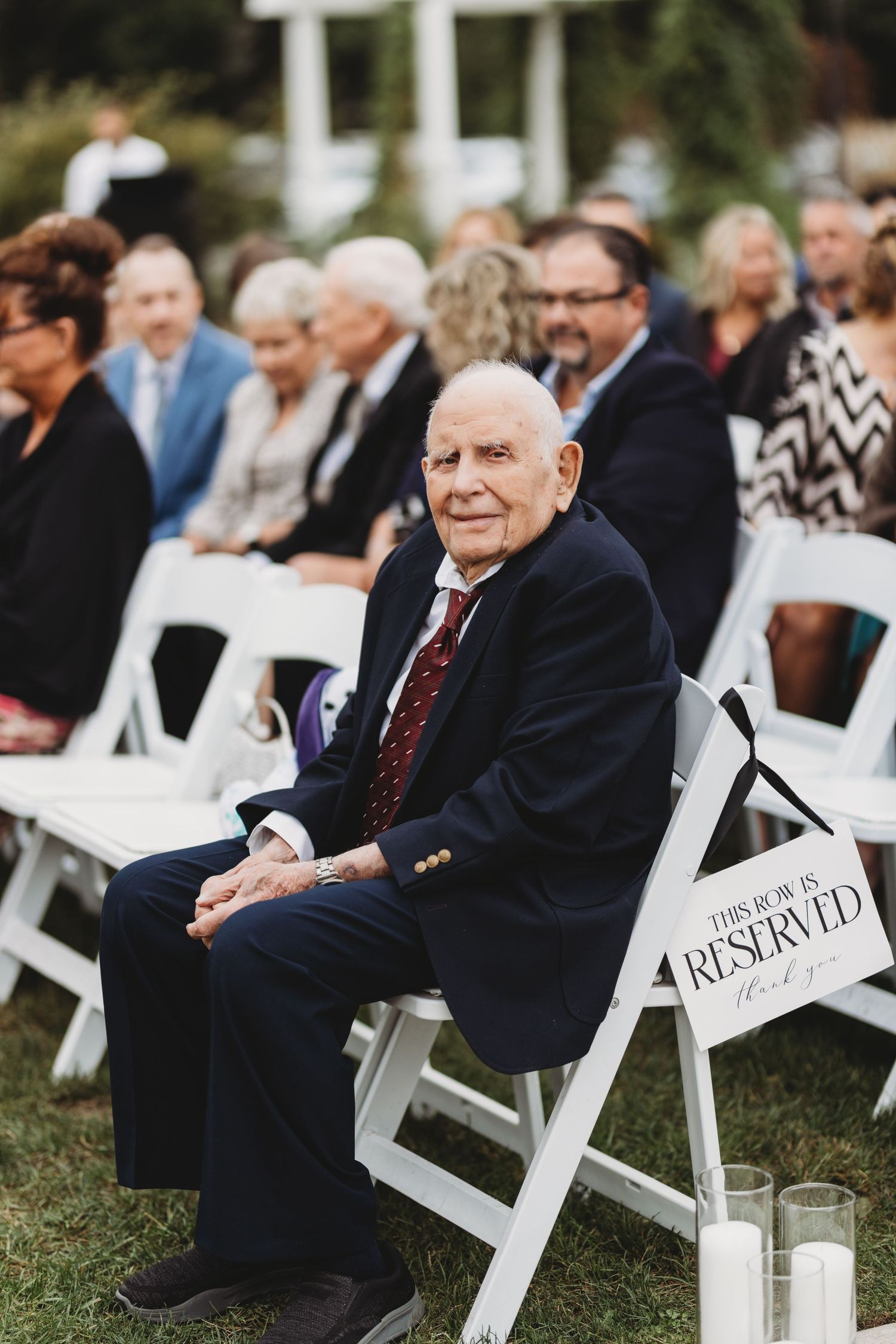 Elderly gentleman in navy suit seated in reserved front row at outdoor wedding ceremony.