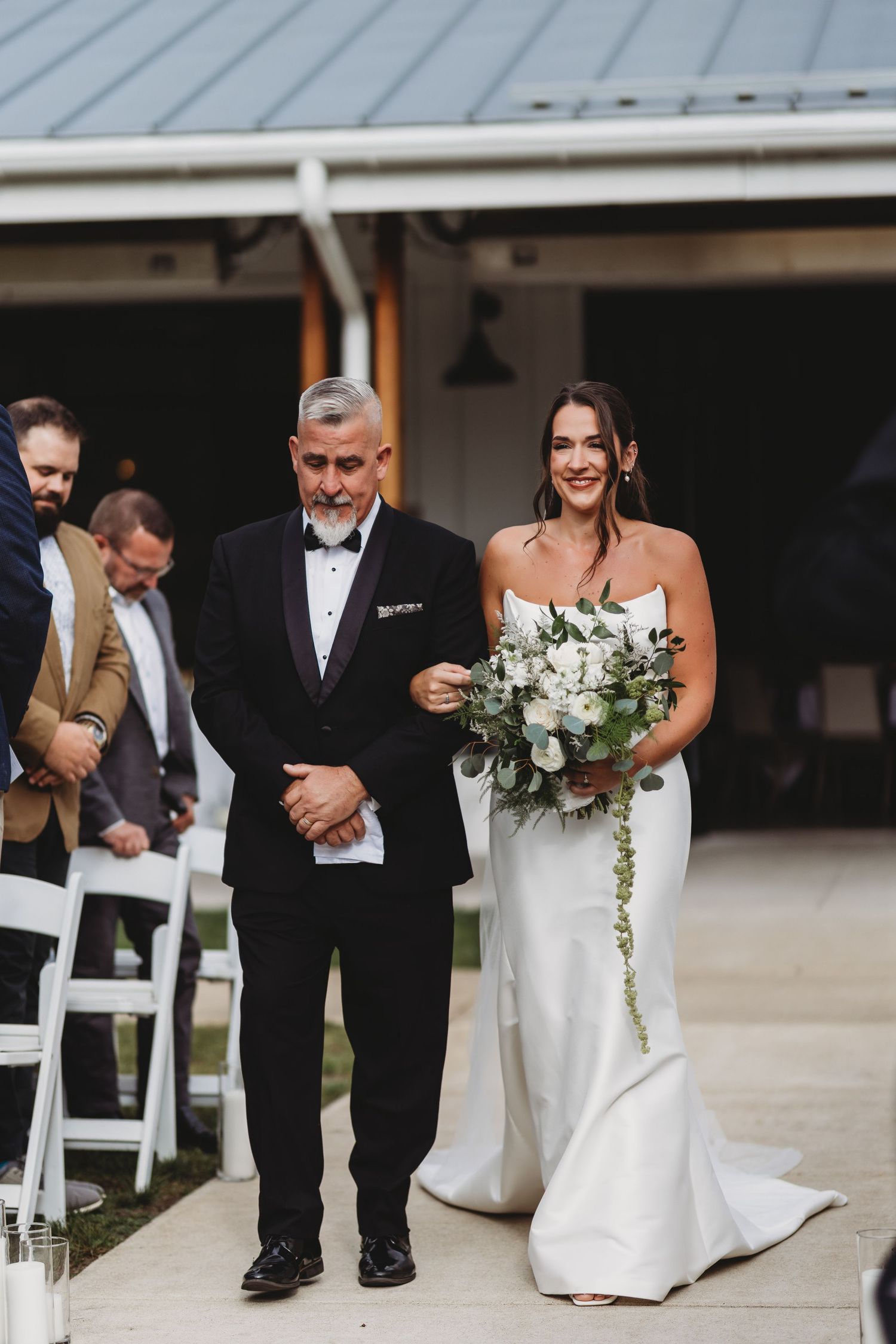 Bride walking down the aisle with her father during outdoor wedding ceremony holding white bouquet.