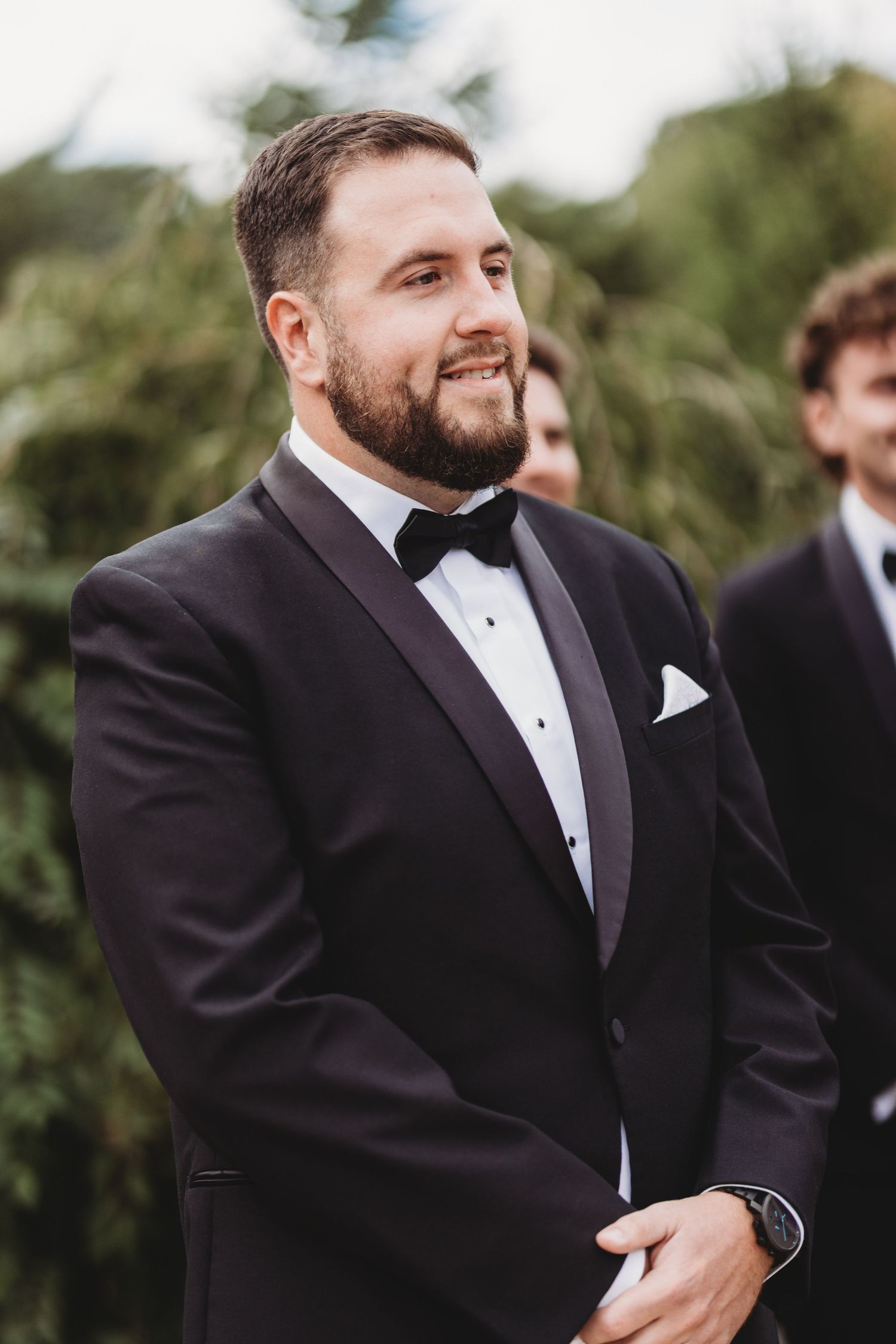 Groom in black tuxedo with bow tie smiling outdoors at wedding ceremony.